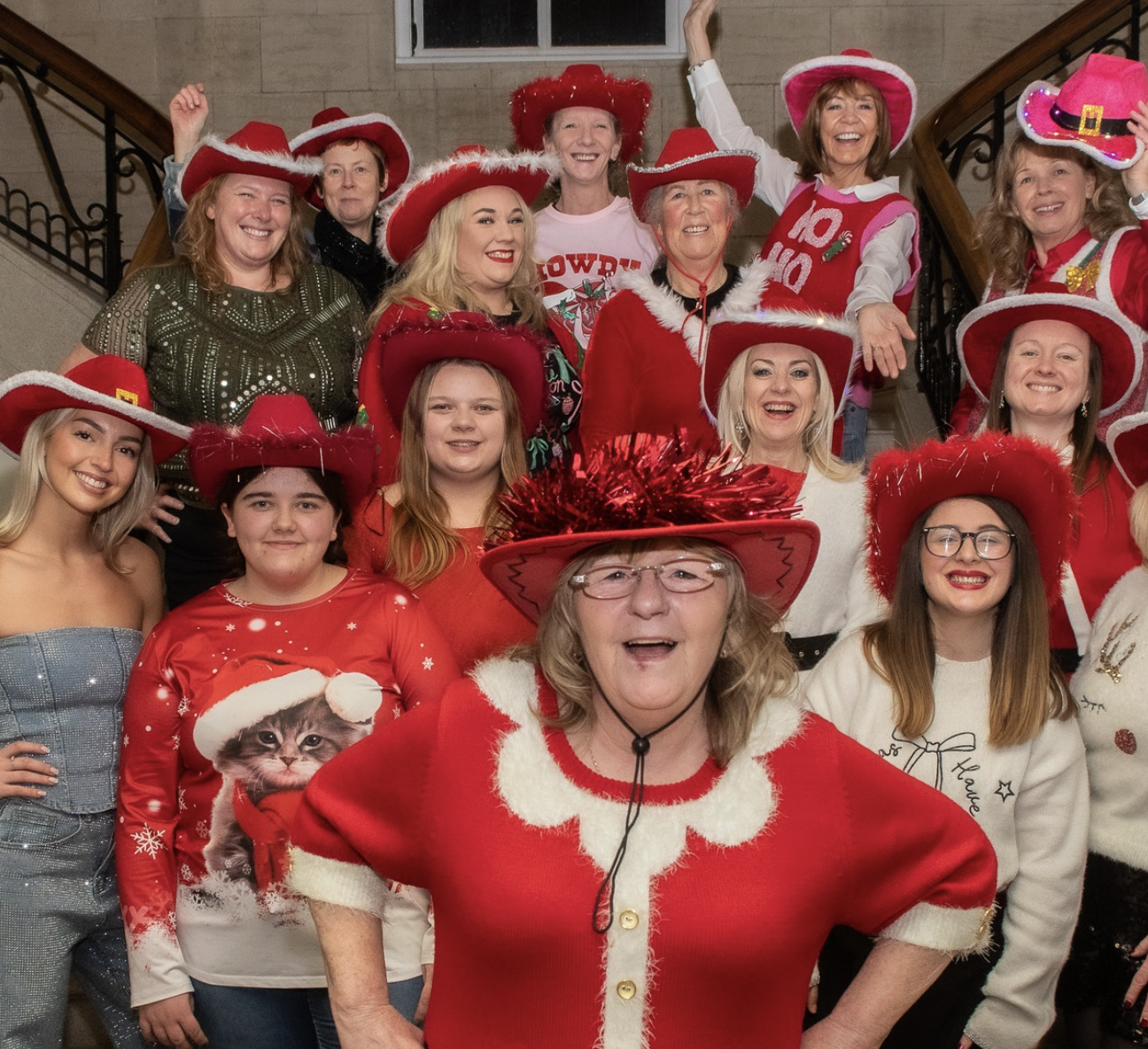 Group of women and girls wearing festive Christmas sweaters and large red hats, smiling for a photo on a staircase indoors.