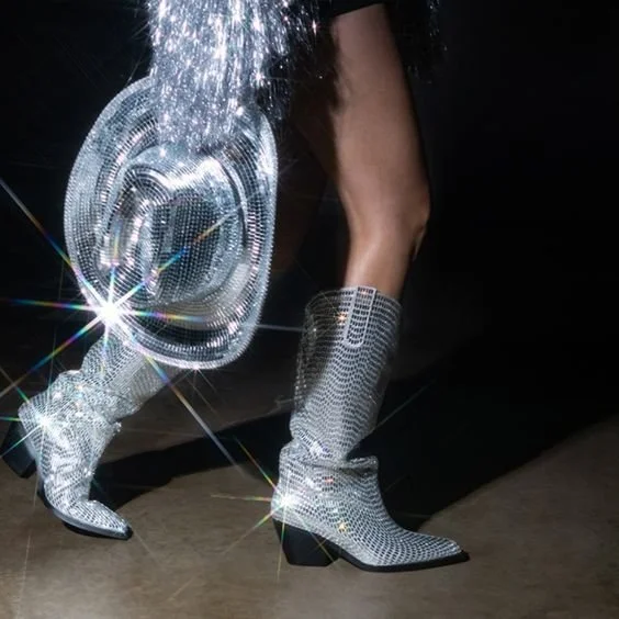 Person walking wearing silver, rhinestone-covered cowboy boots, holding a matching rhinestone-covered purse, and wearing a fringed silver outfit.