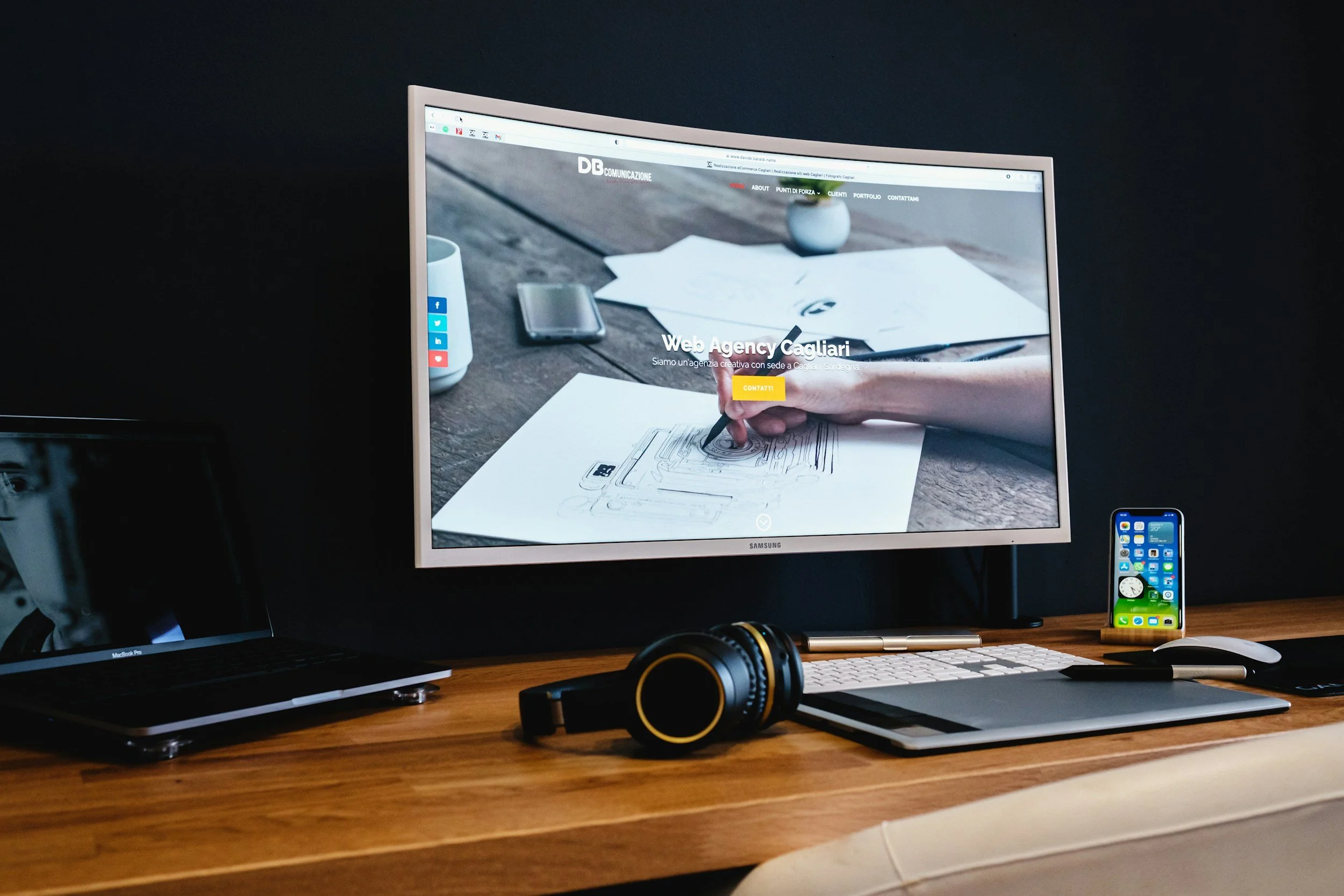 A monitor on a desk opened to a website with headphones, keyboard, mouse and cellphone surrounding it.