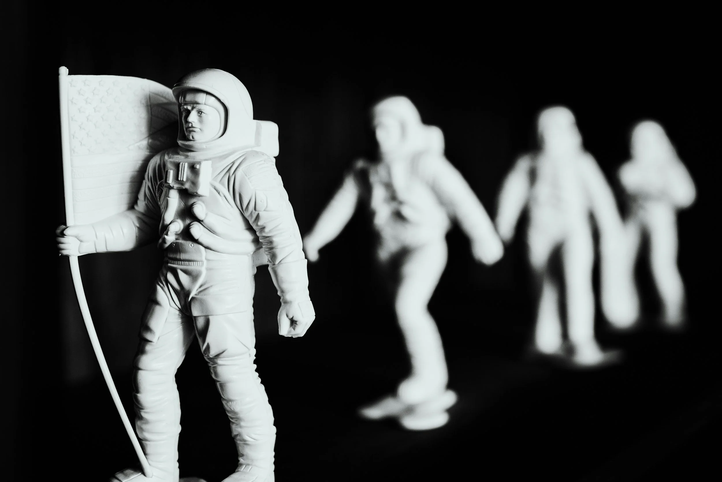 Black and white photo of four astronaut figurines in a line, with the first in focus holding an American flag.