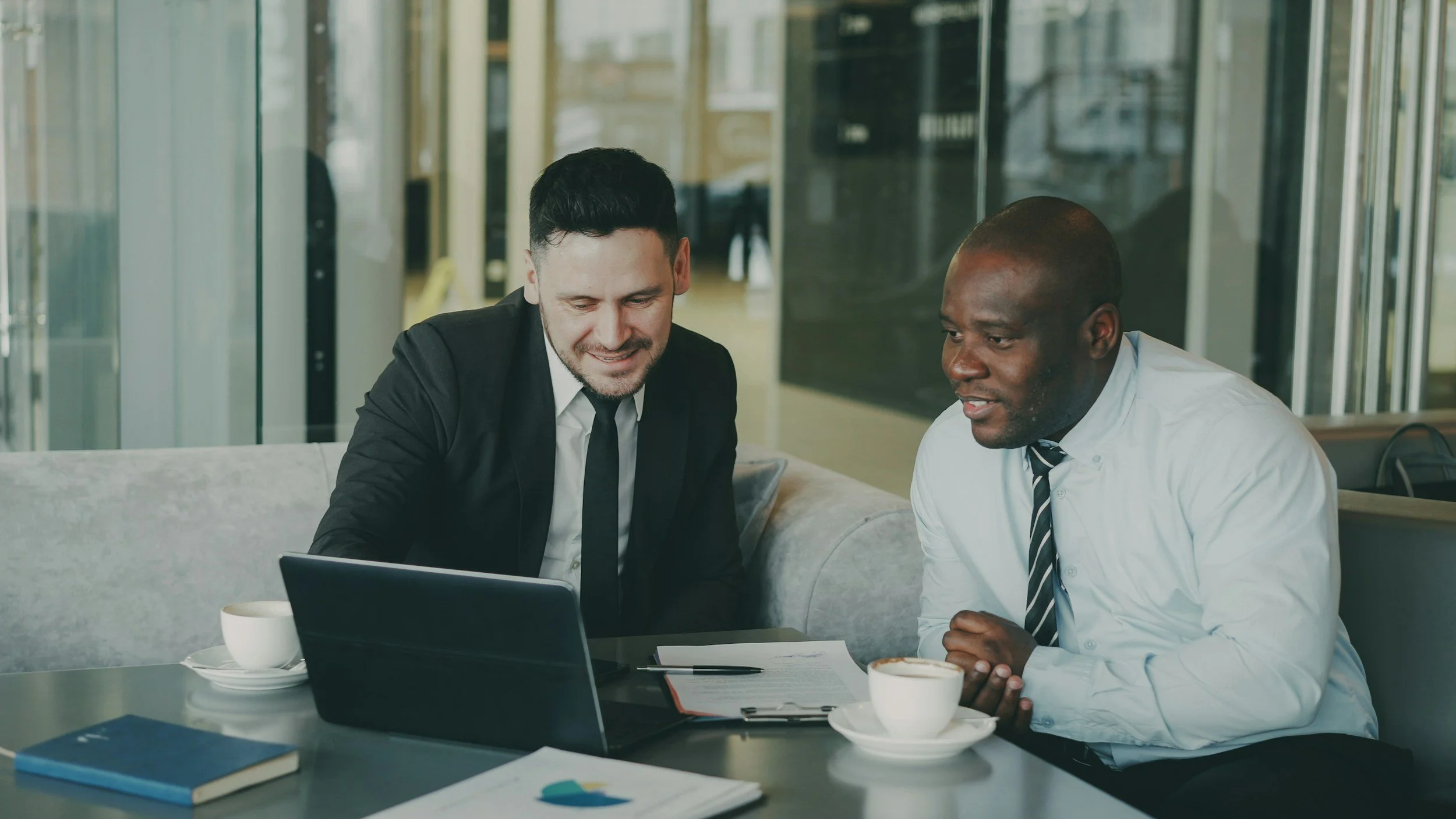 Two men in business attire sitting at a table, looking at a laptop and discussing, with notes and coffee cups in front of them, in a modern office or cafe setting.