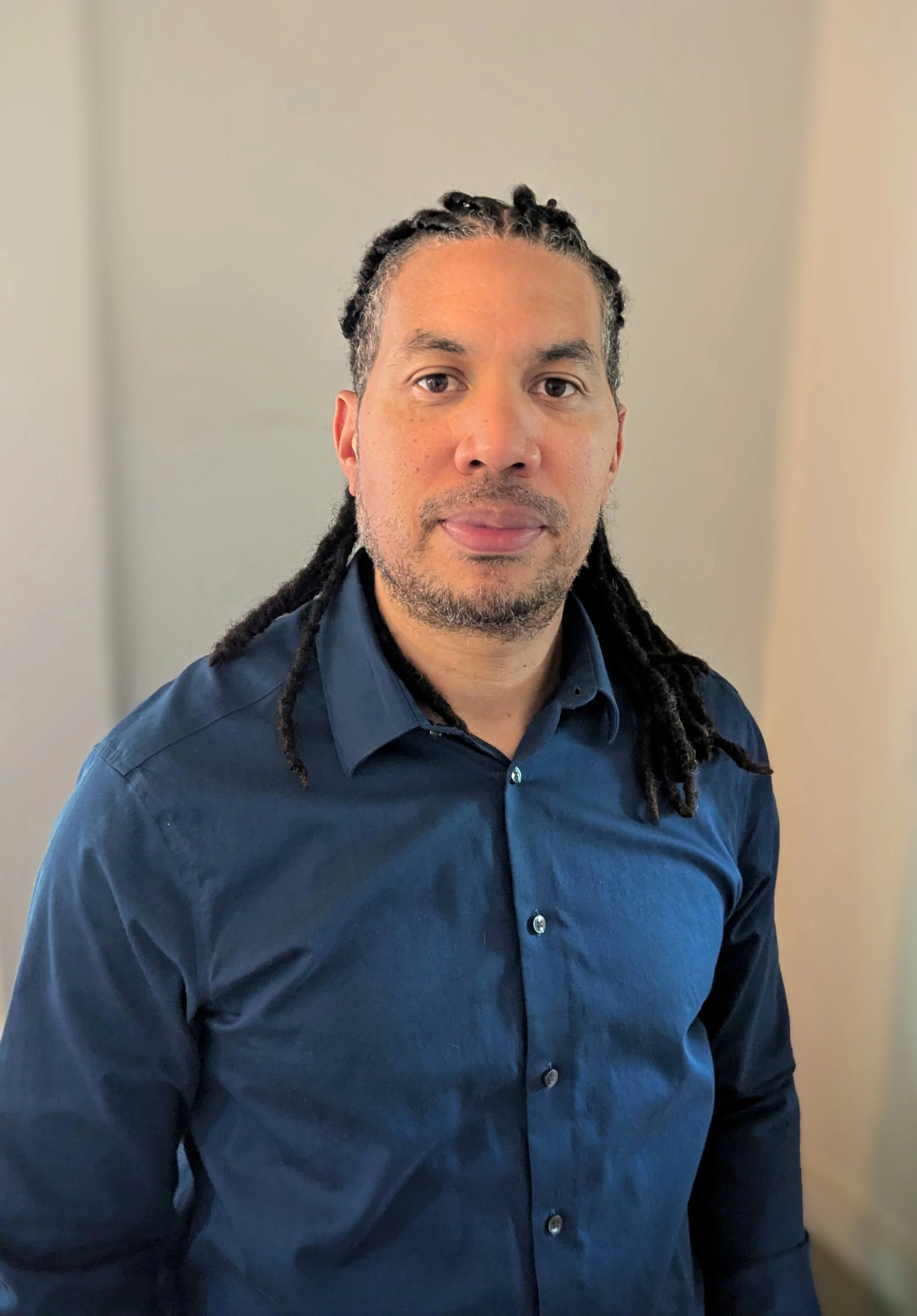 A man with dark hair and dreadlocks wearing a dark blue button-up shirt, standing indoors in front of a plain wall.