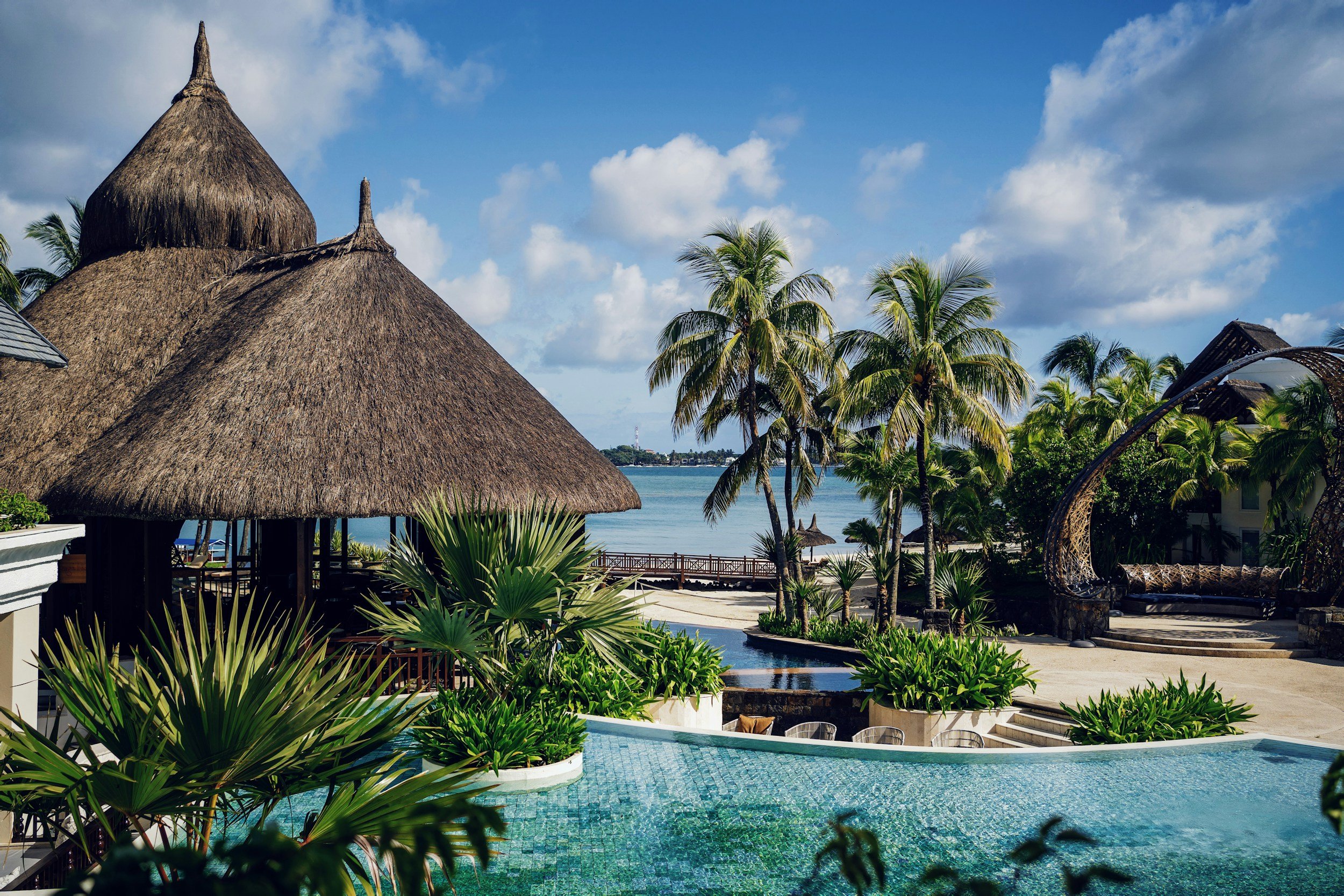 Tropical resort with thatched huts, palm trees, a swimming pool, and a view of the ocean under a partly cloudy sky.