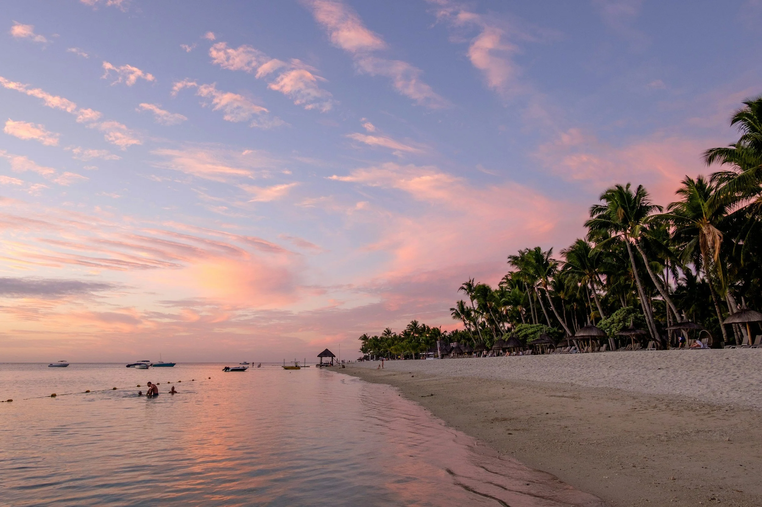 A tropical beach during sunset with colorful sky, palm trees lining the shore, thatched umbrellas and lounge chairs on the sand, and boats floating on the calm water.