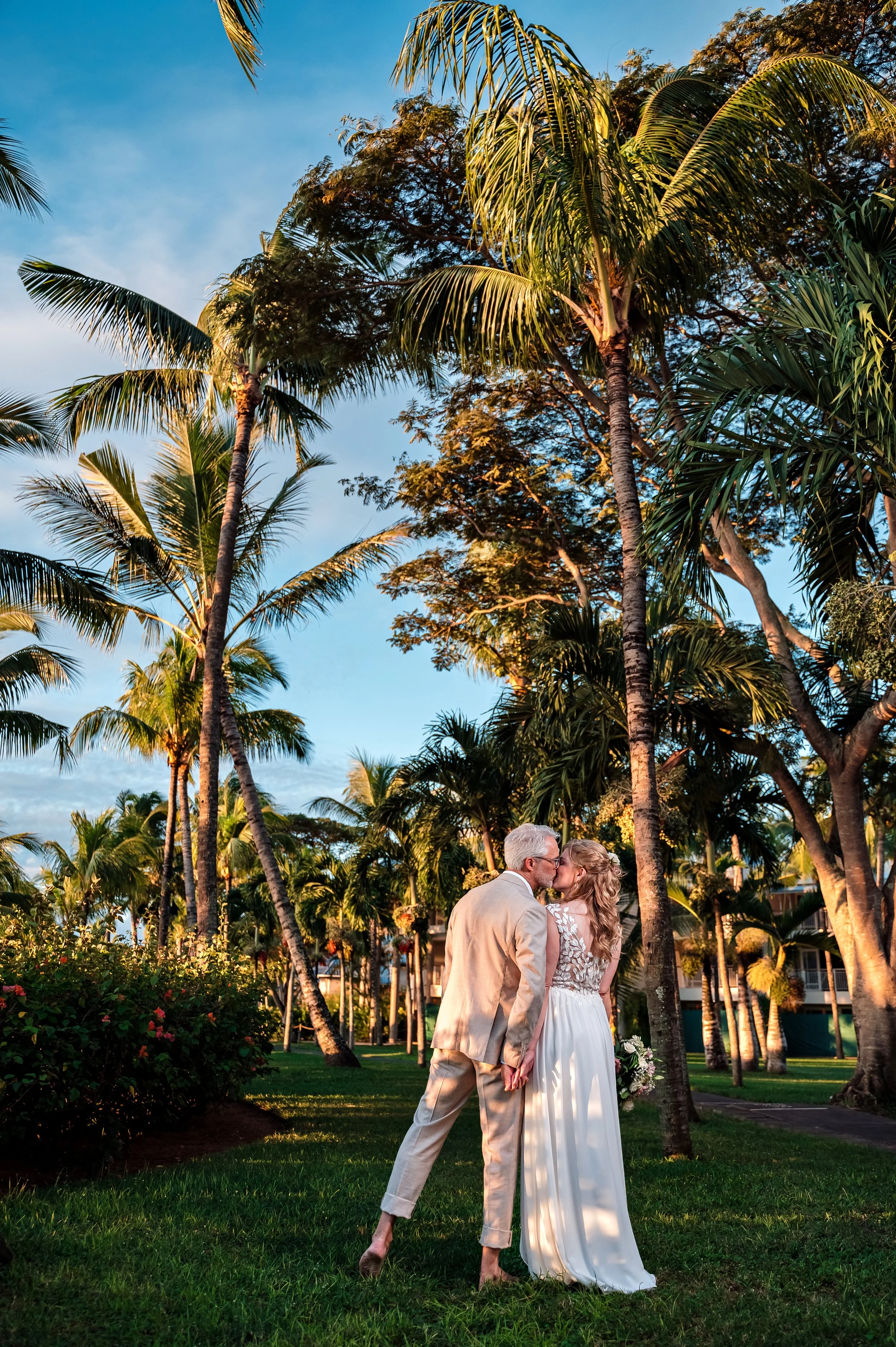 Bride and groom under tall palm trees and green grass at sunset.