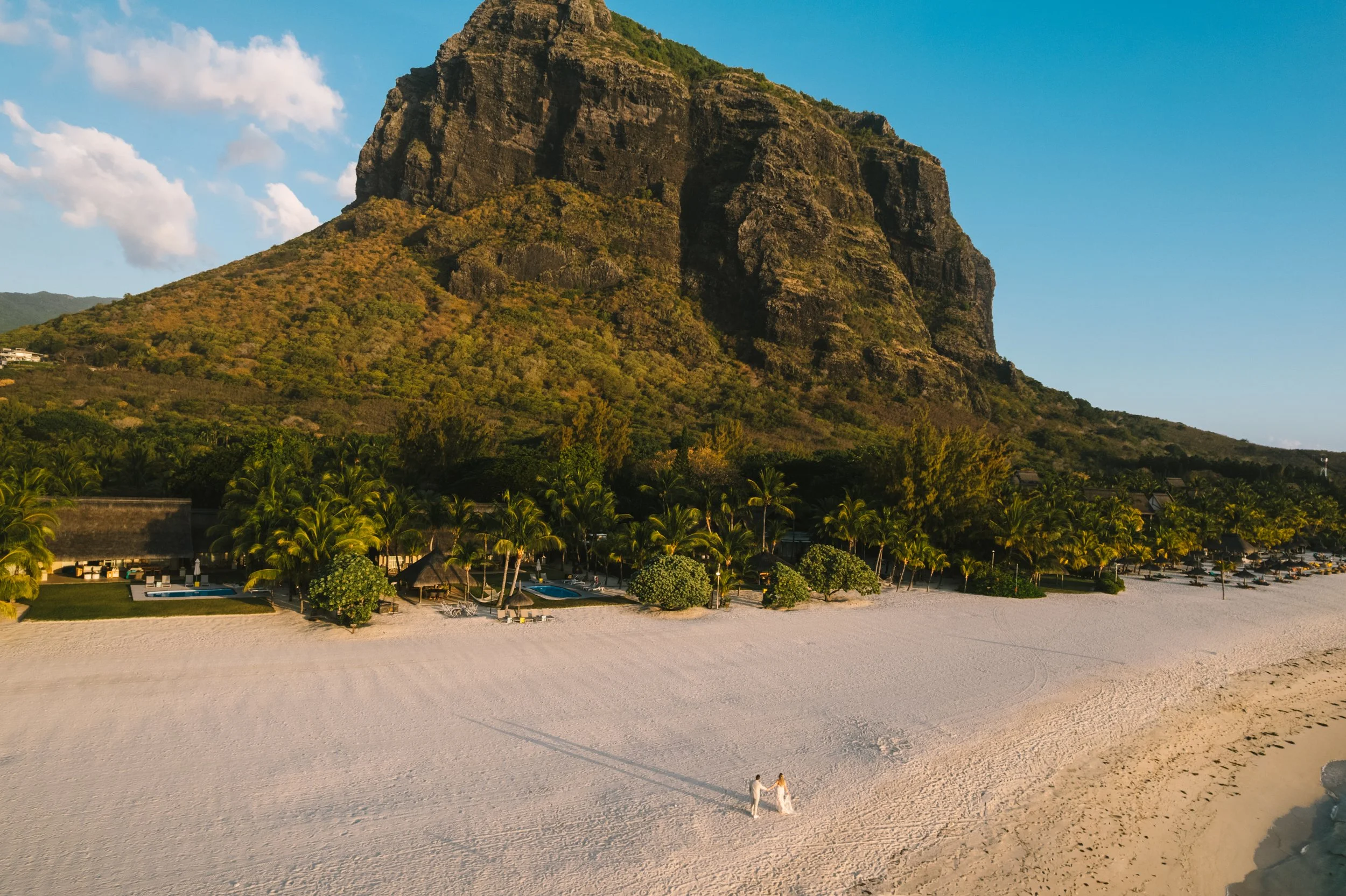 A couple in wedding attire holding hands on a sandy beach with palm trees, thatched-roof cabanas, and a large rocky mountain in the background under a blue sky.