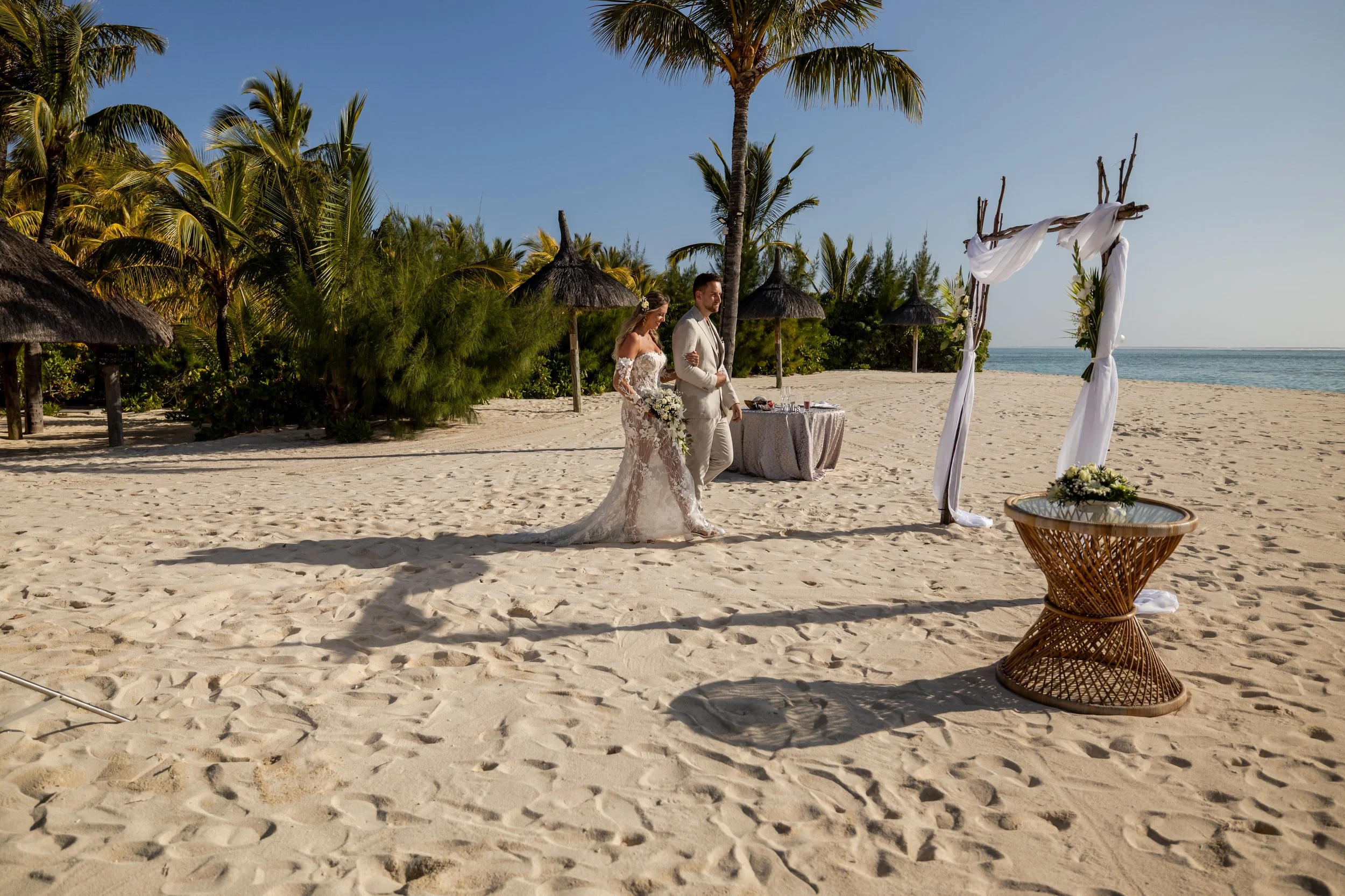 A couple getting married on a beach with palm trees, a wooden arch draped with white fabric, and a table with a flower arrangement.
