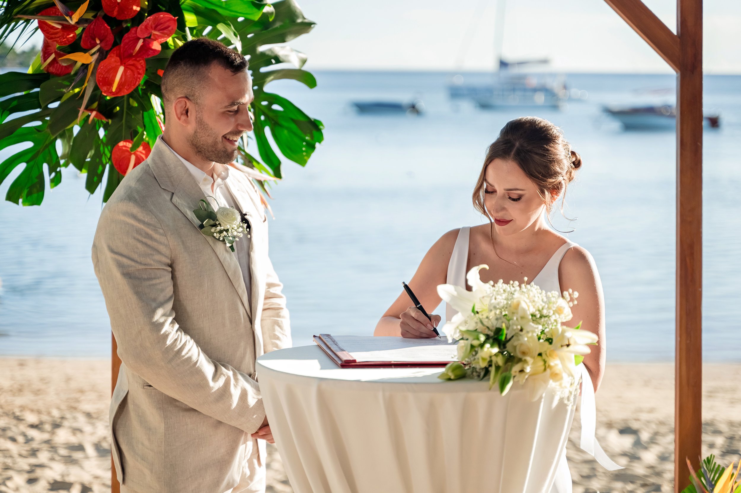 A bride and groom signing wedding documents at a beachside wedding altar with boats in the background. The bride is wearing a white dress and the groom is in a beige suit.
