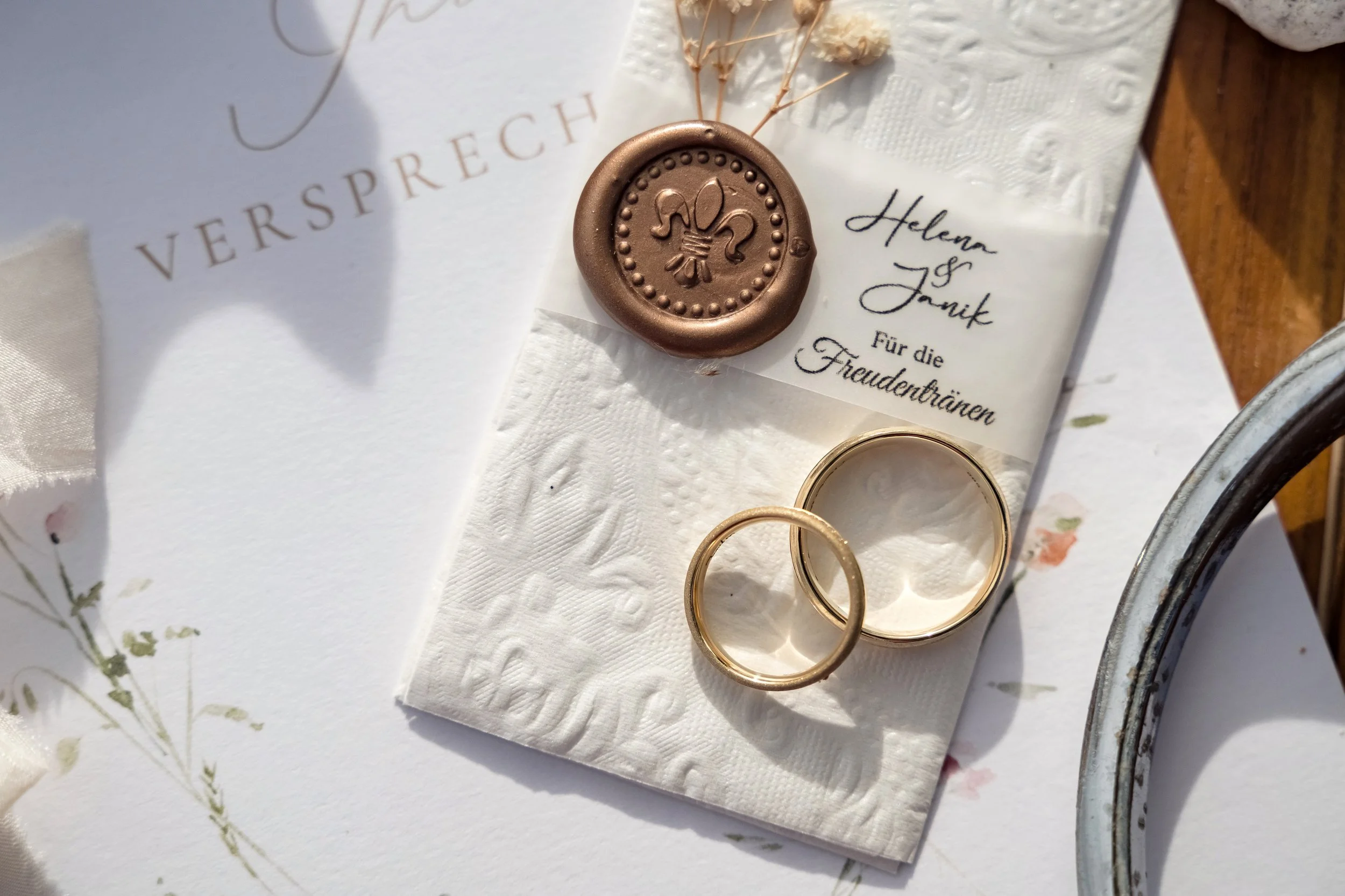 Close-up of a wedding ring set on a white napkin with embossed patterns, two gold rings and a large wedding band, with a white floral envelope in the background, and a brown wax seal with a fleur-de-lis design on top of the napkin.