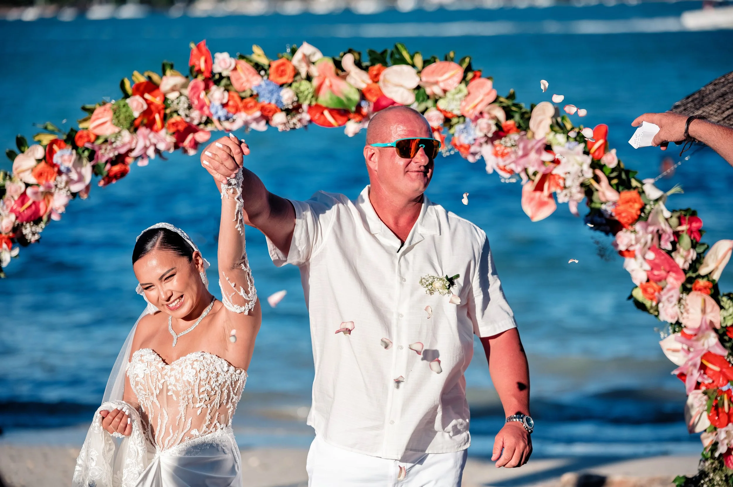A couple getting married on the beach, with the groom wearing sunglasses and the bride smiling, walking under a floral archway, with the ocean in the background.