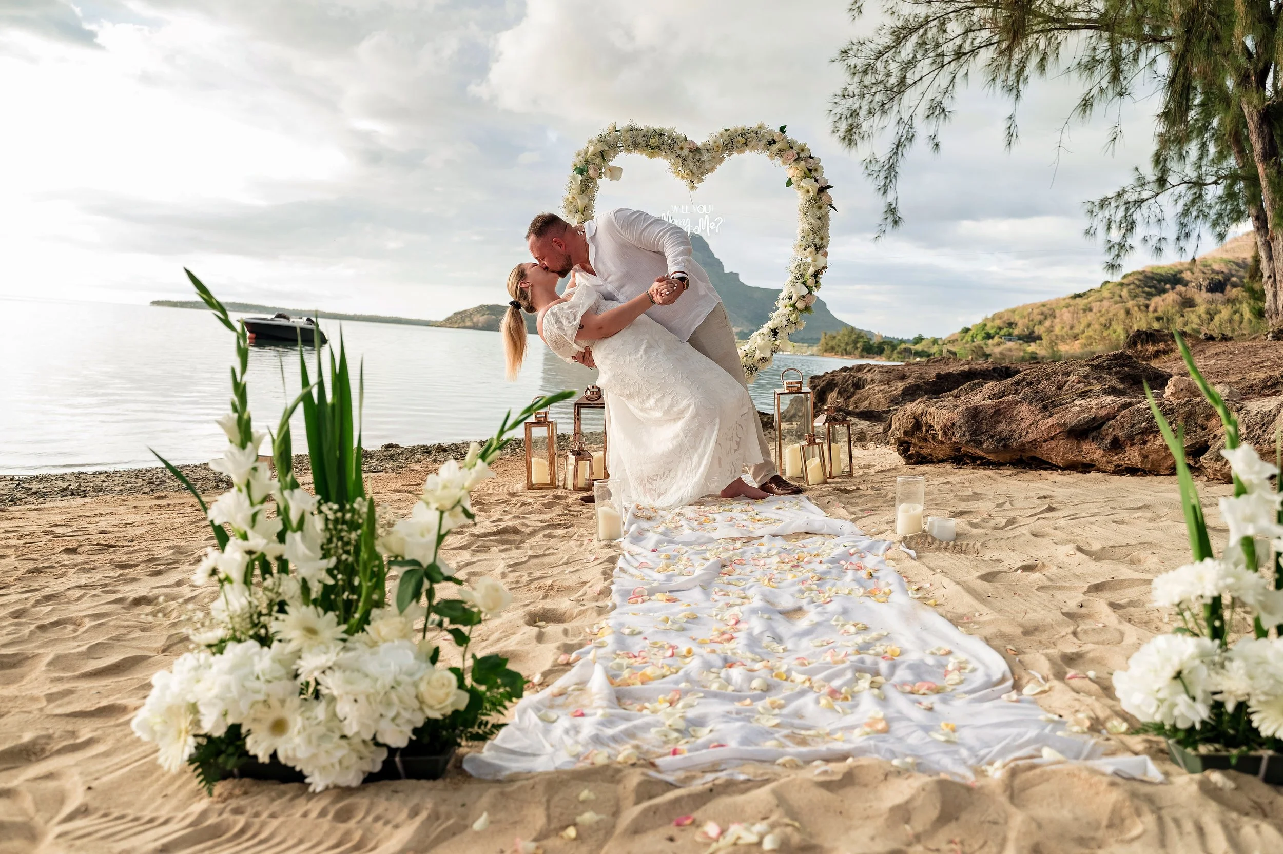 A couple shares a kiss during a beach wedding at sunset, with a floral heart backdrop, candles, and flower arrangements on the sand.
