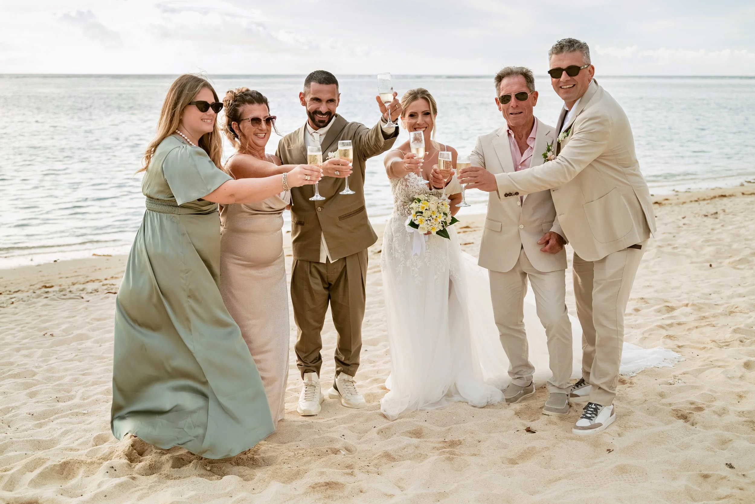 Group of people in wedding attire celebrating with champagne on a beach, with ocean in the background
