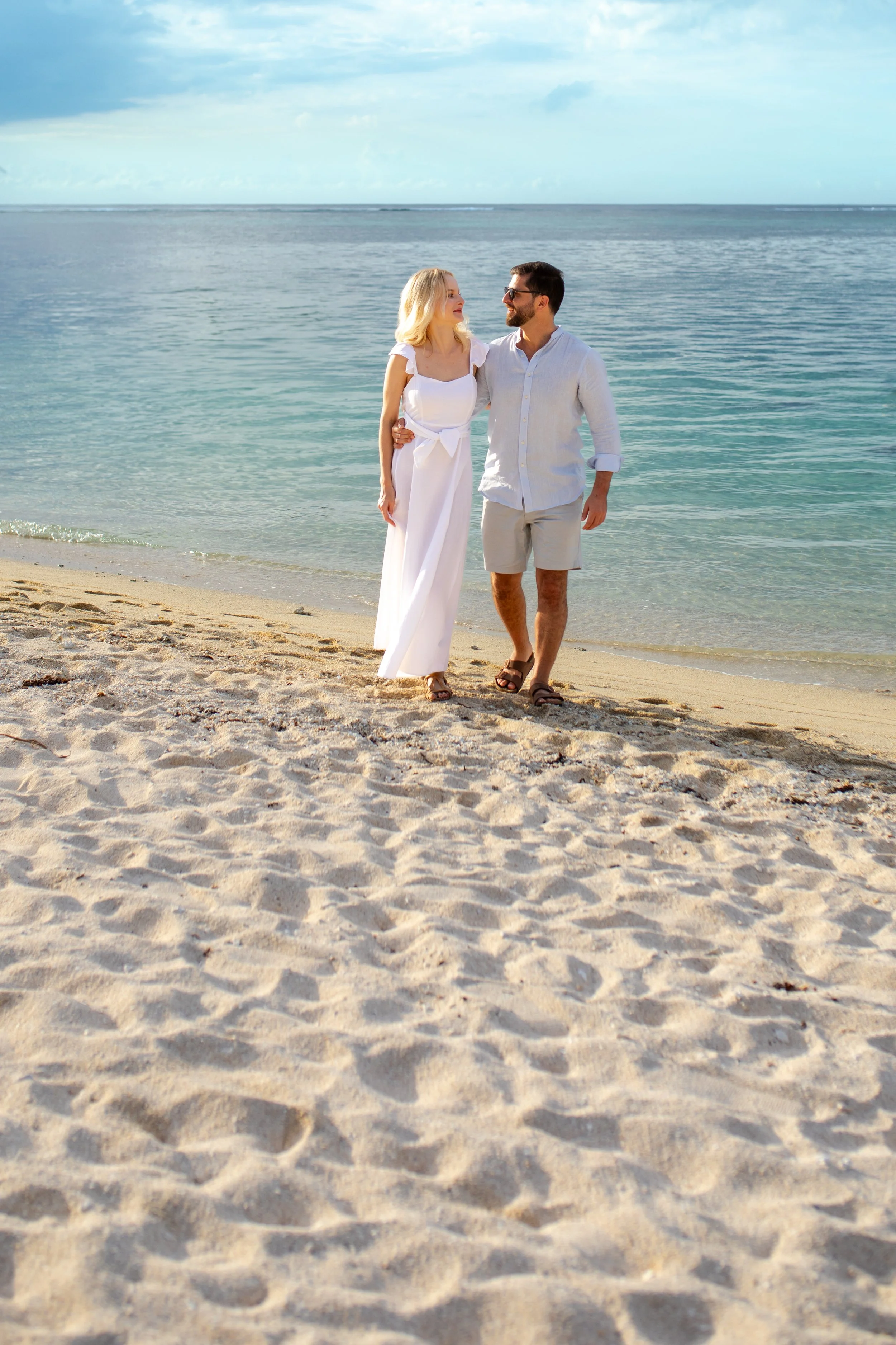 A couple walks on the beach near the ocean, with the woman wearing a white dress and the man in a white shirt and shorts.