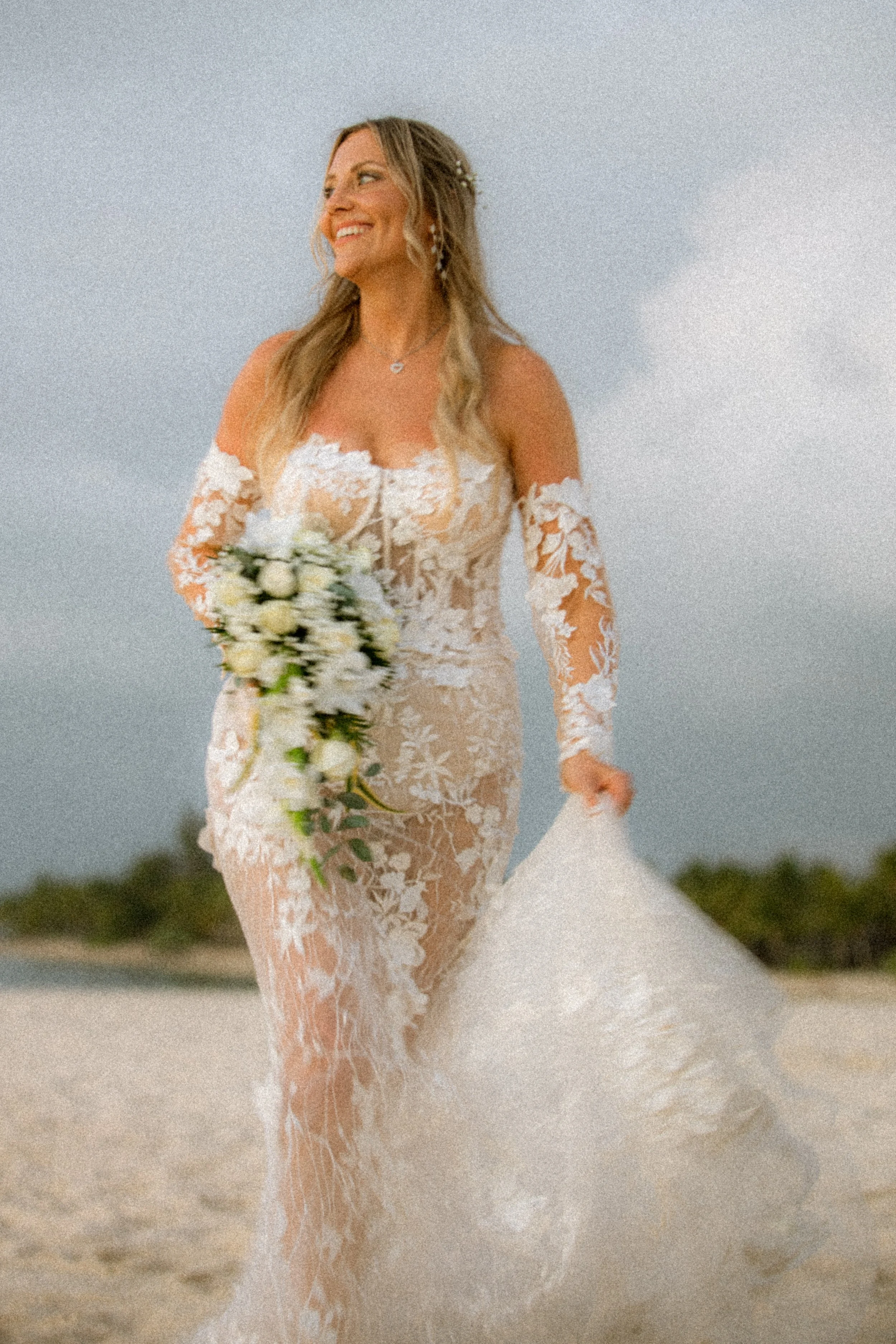 A smiling bride in a sheer lace wedding dress holding a bouquet of white flowers on the beach.