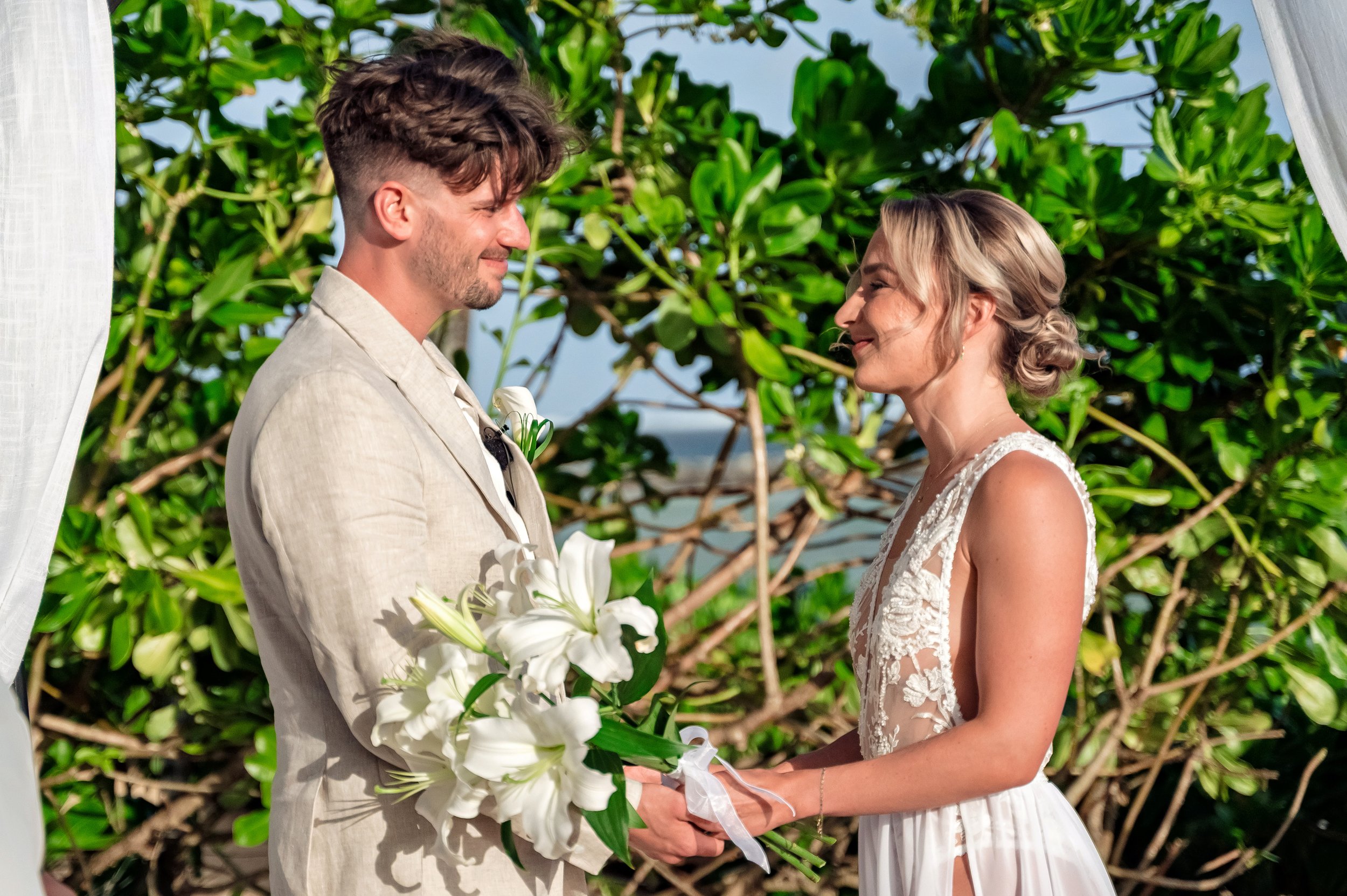 A groom and bride standing facing each other outdoors during a wedding ceremony, with the groom holding a bouquet of white lilies, surrounded by green foliage.