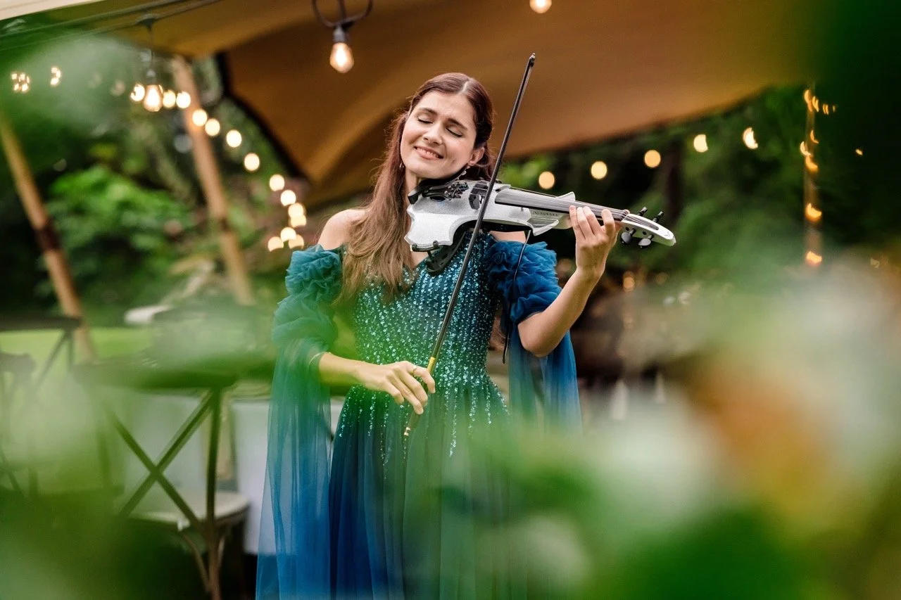 A woman in a blue, sequined dress playing a violin outdoors during a celebration or event, with blurred greenery and string lights in the background.