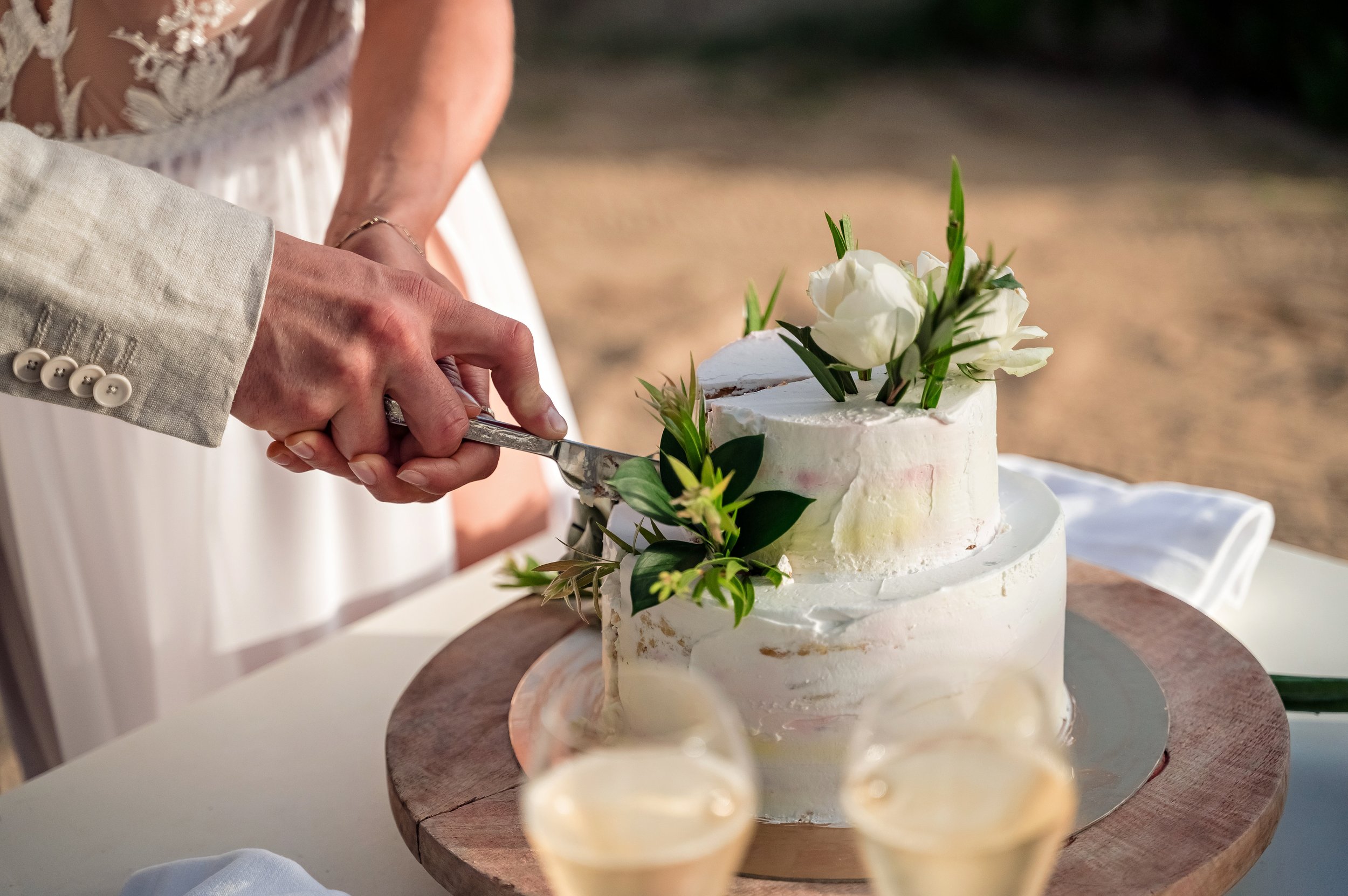 A couple's hands cutting a wedding cake decorated with white flowers and greenery