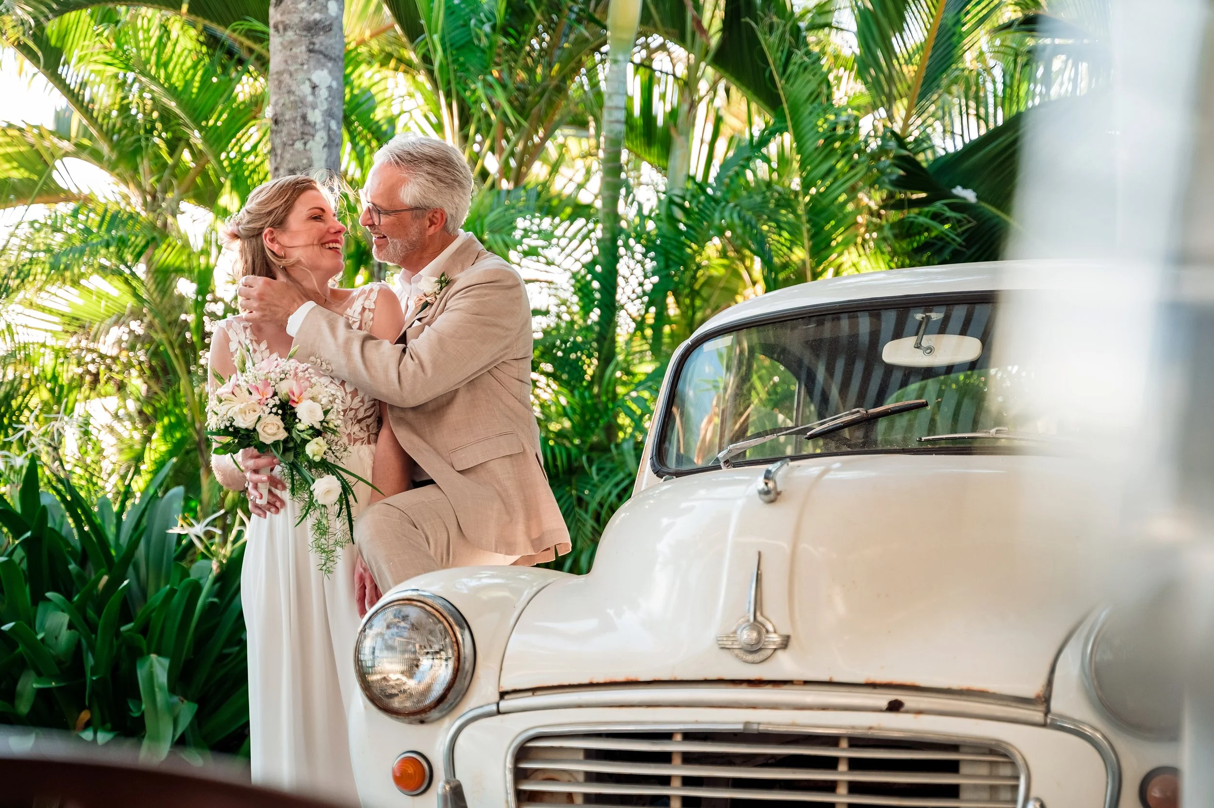 A couple in wedding attire celebrating outdoors among lush green plants, embracing each other while standing next to a vintage white car.