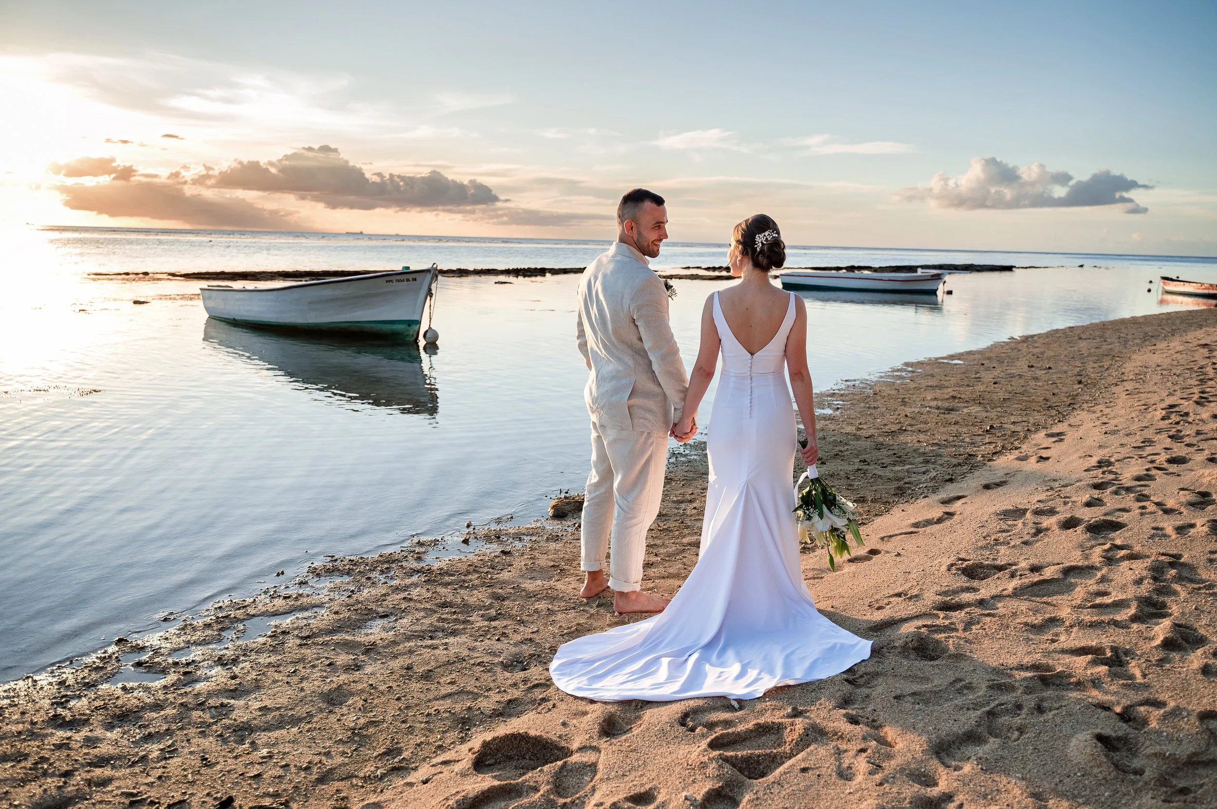 A newlywed couple holding hands on a beach during sunset, with boats floating on calm water in the background.