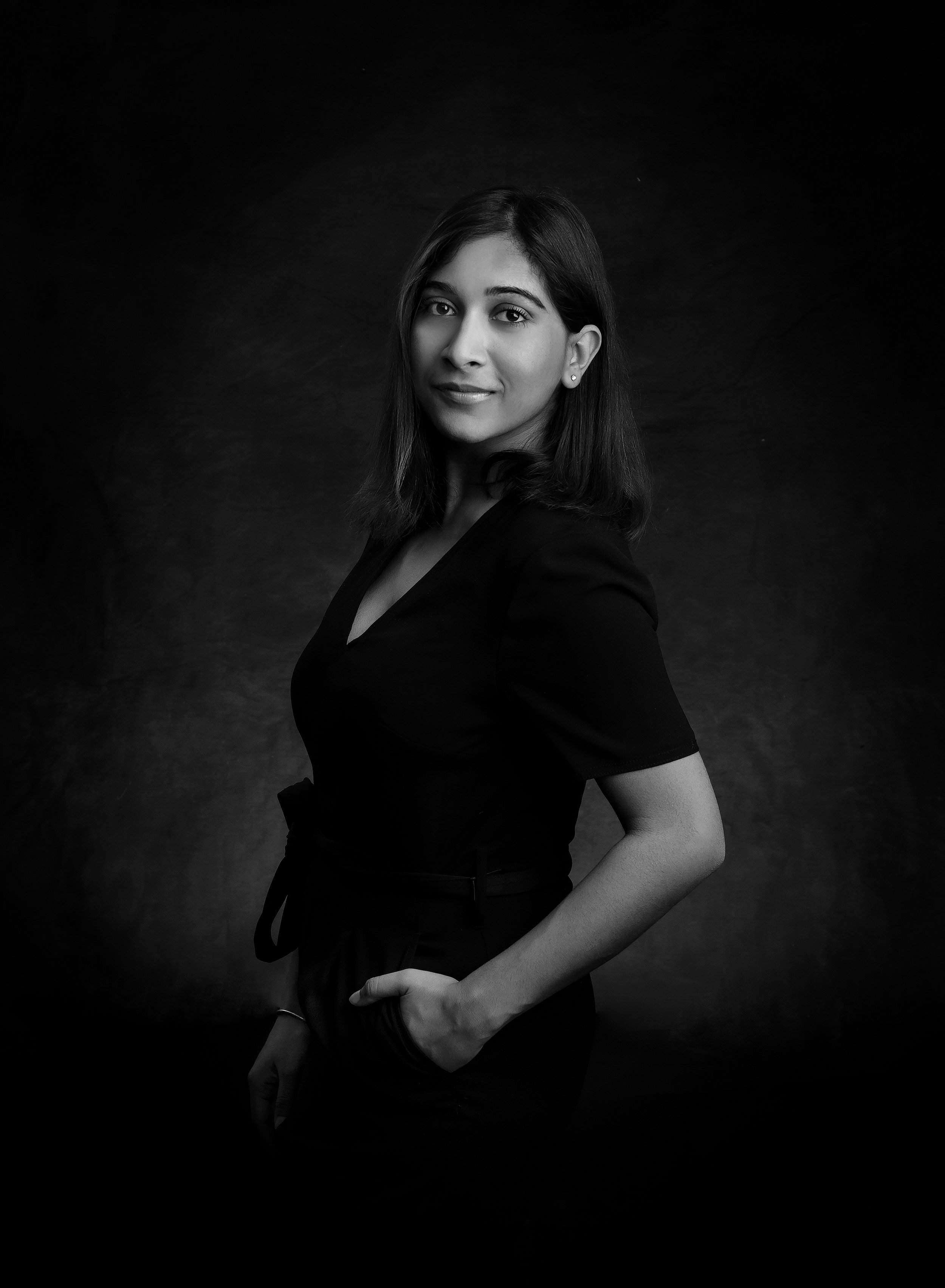Black and white portrait of a young woman with shoulder-length hair, wearing a dark top with short sleeves, standing against a dark background.
