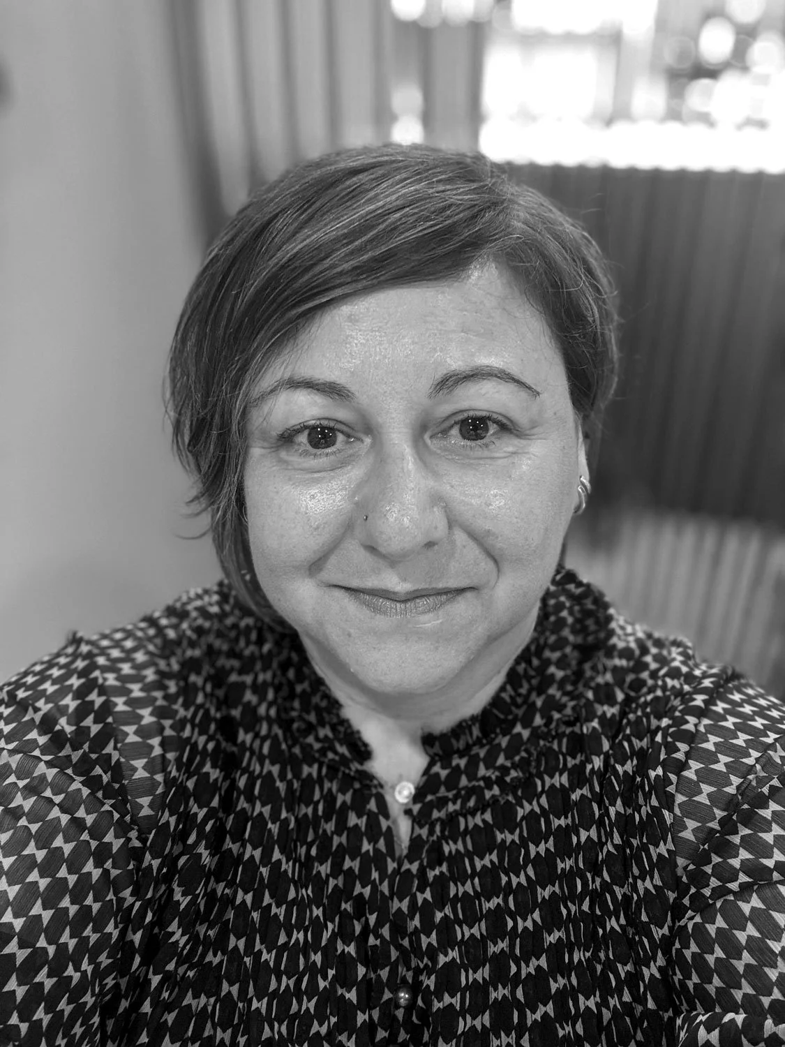 A woman with short hair and a gentle smile, wearing a patterned blouse, in an indoor setting with wooden paneling and windows in the background.