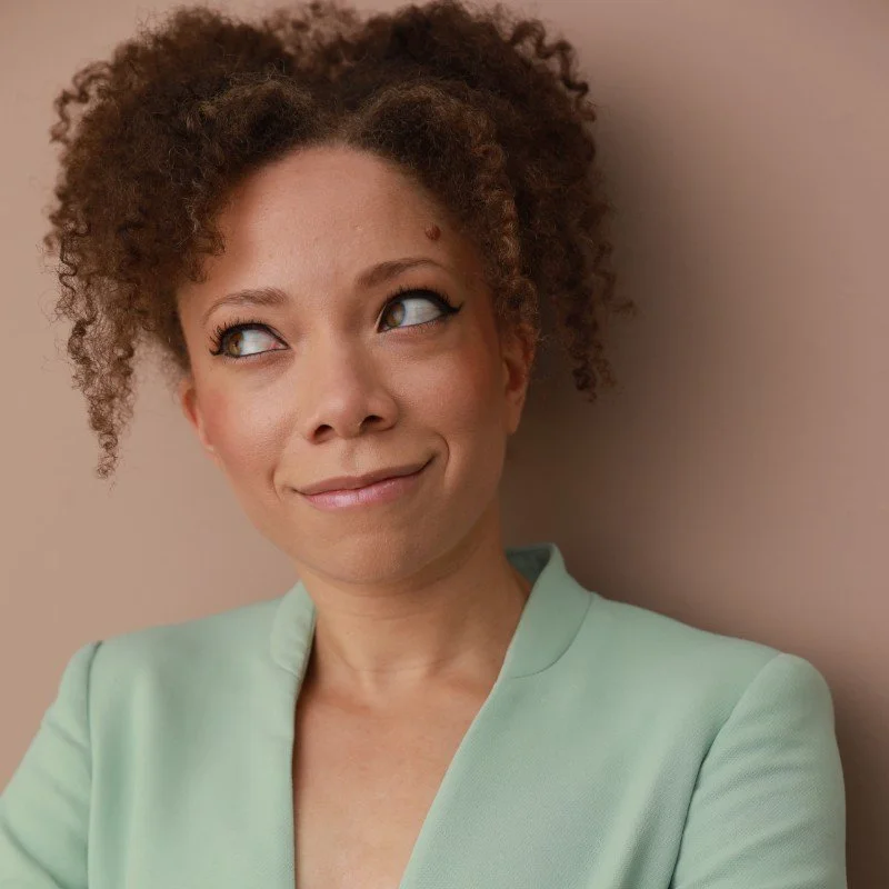 A woman with curly hair, wearing a light green blazer, smiling thoughtfully, standing against a beige wall.