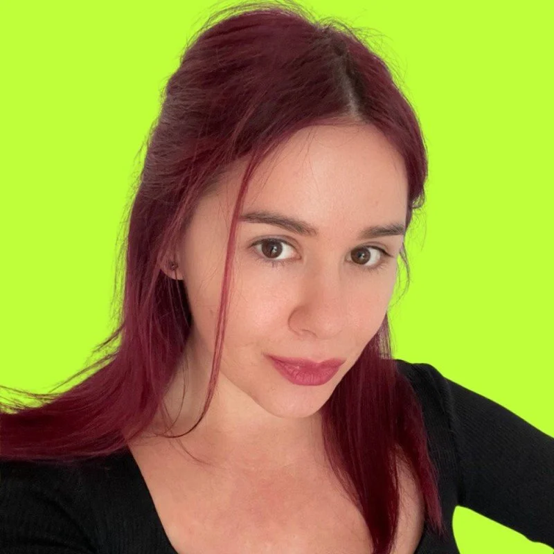 A young woman with shoulder-length red hair and fair skin takes a selfie against a bright green background, wearing a black top.