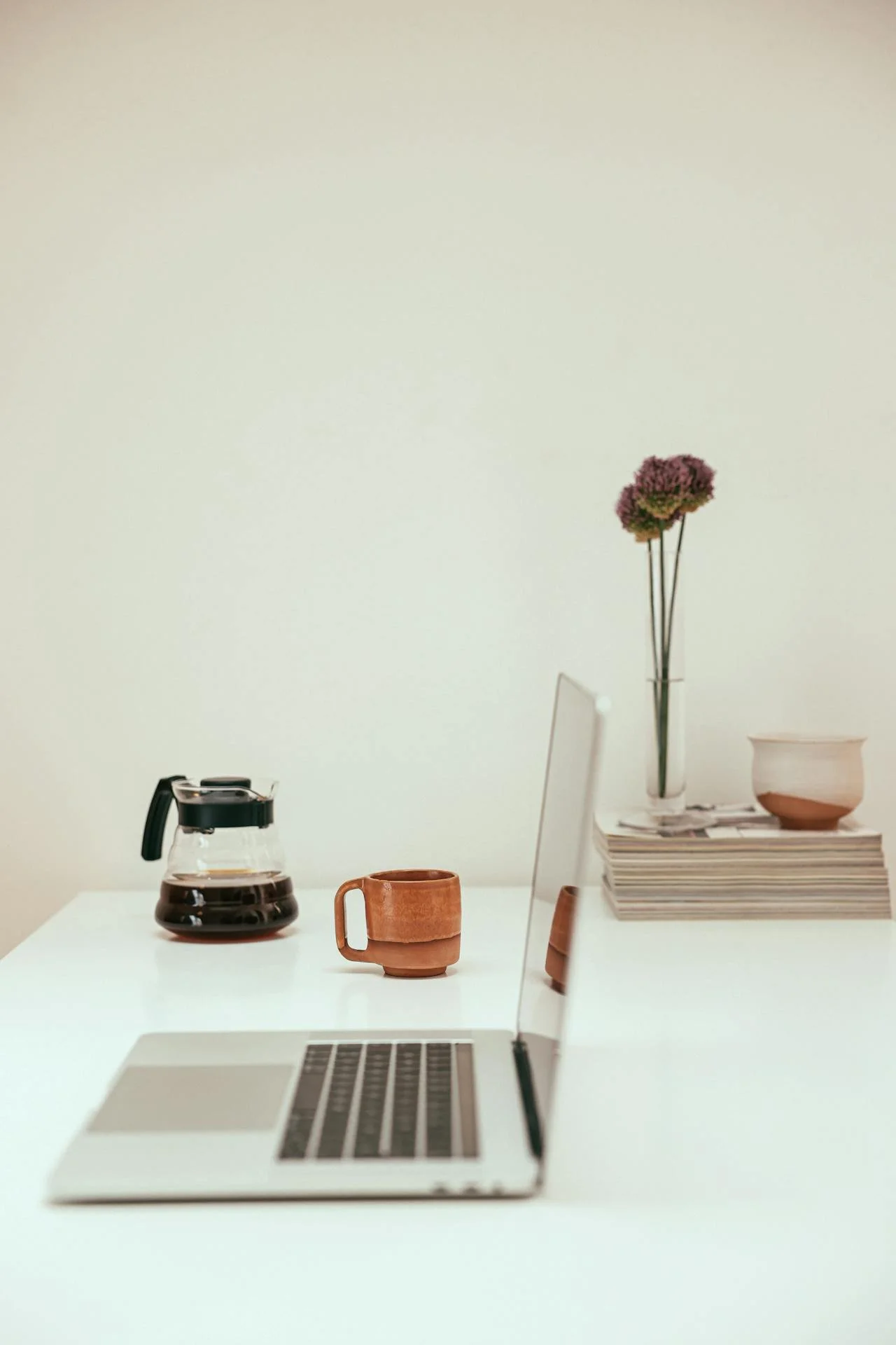 A minimalist workspace with a silver laptop, a terracotta mug, a glass coffee pot, a pink flower in a tall glass vase, a small bowl, and a stack of magazines on a white table against a plain light-colored wall.