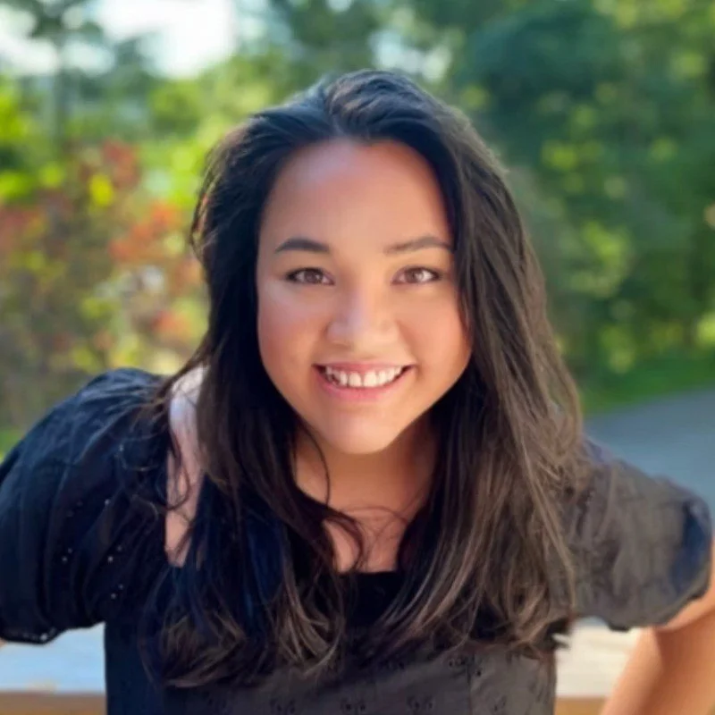 A young woman smiling outdoors with green trees in the background.