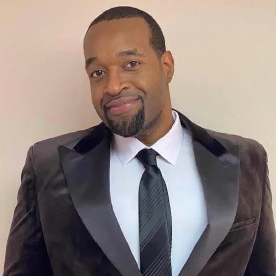 A man in a formal black tuxedo with satin lapels, white dress shirt, and black tie, standing against a plain light-colored background. He has short hair, a goatee, and a slight smile.