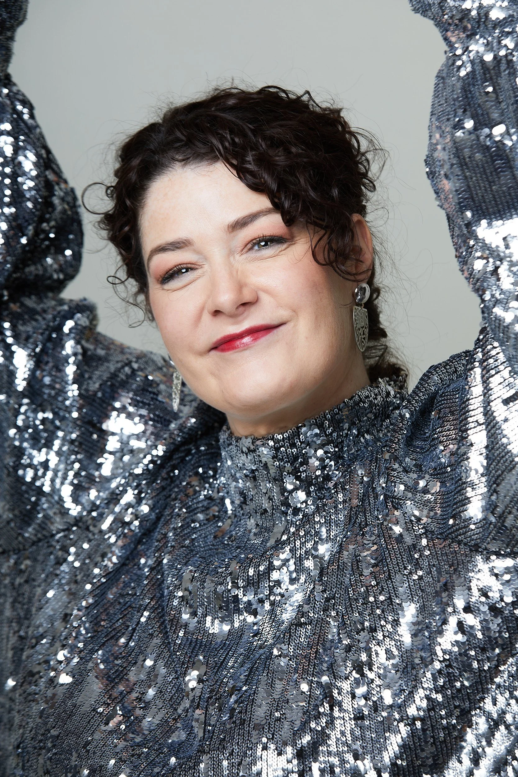 Woman with short curly dark hair winks and smiles, wearing a shiny silver sequin top and silver earrings.