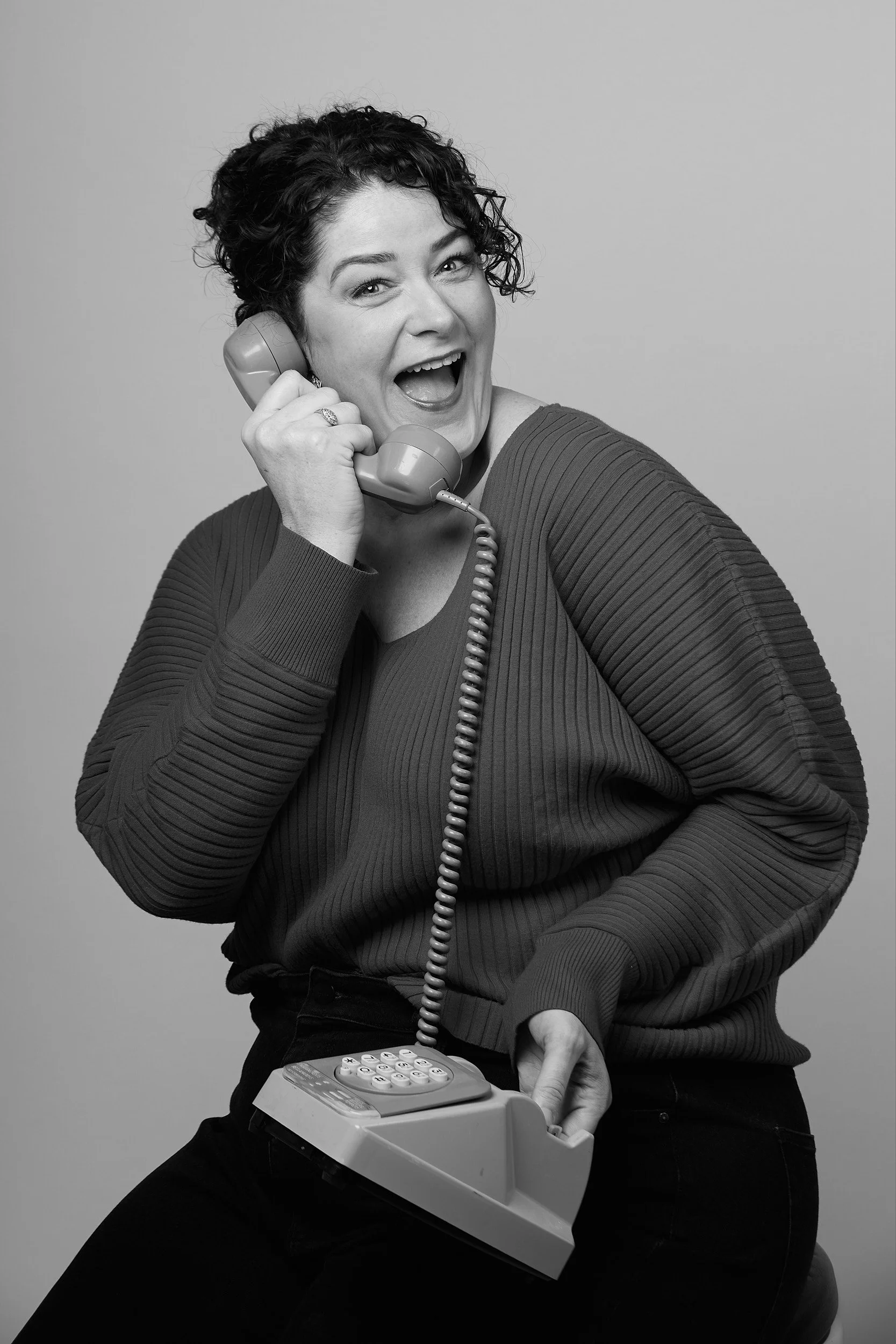 A woman with short dark curly hair smiling and laughing while holding a vintage corded telephone receiver to her ear. She is wearing a ribbed sweater and dark pants, with the telephone's base resting on her lap.