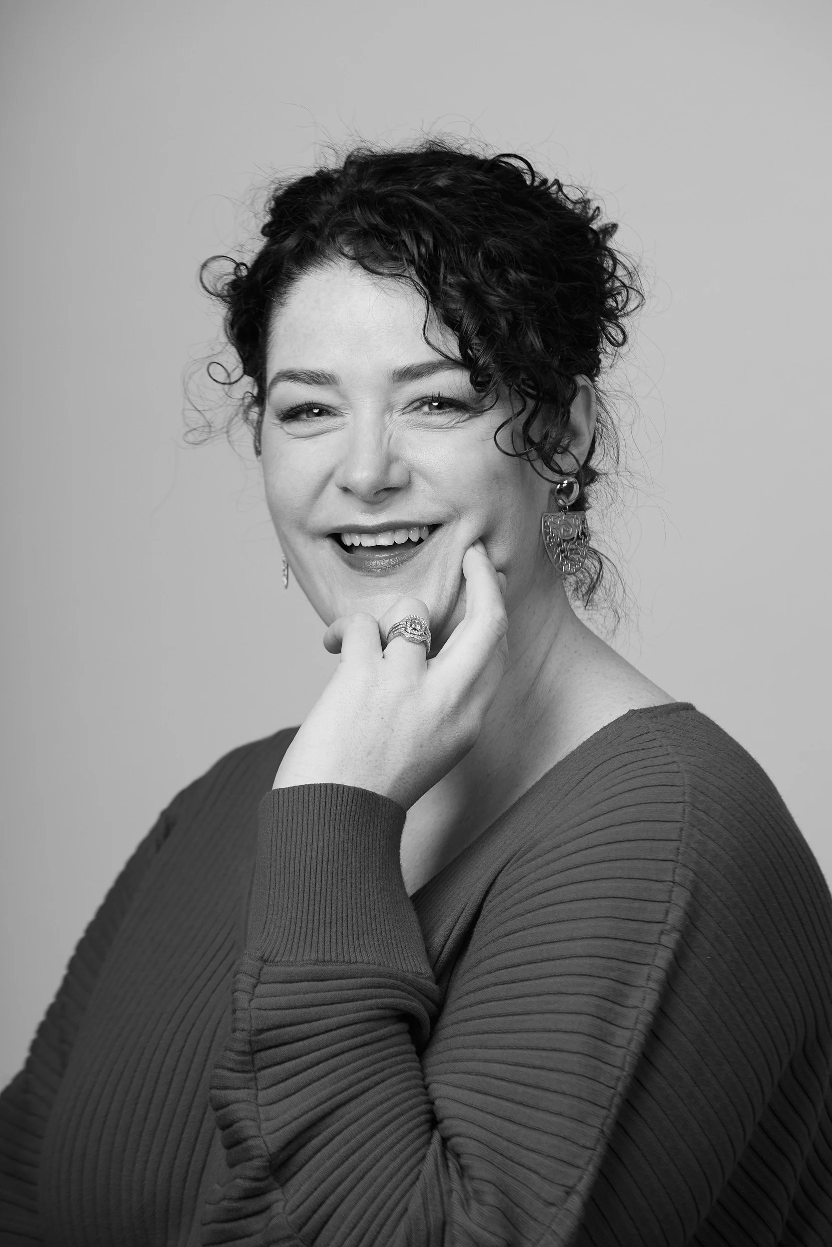 Black and white portrait of a woman with curly hair, smiling and touching her chin, wearing earrings, a ring, and a ribbed long-sleeve top.