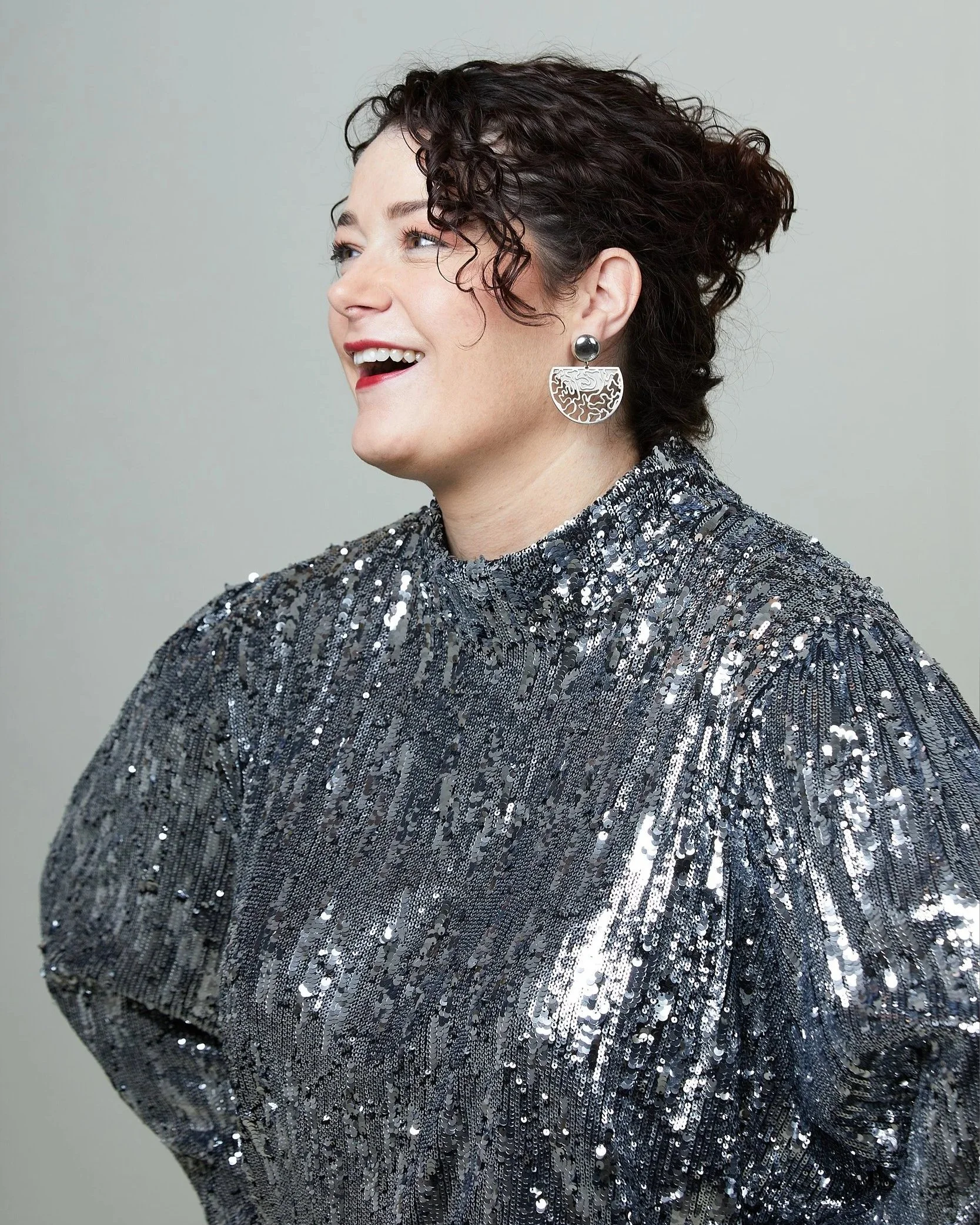 A woman with short curly dark hair wearing large silver earrings and a shiny, sequined silver dress, smiling and looking to her left.
