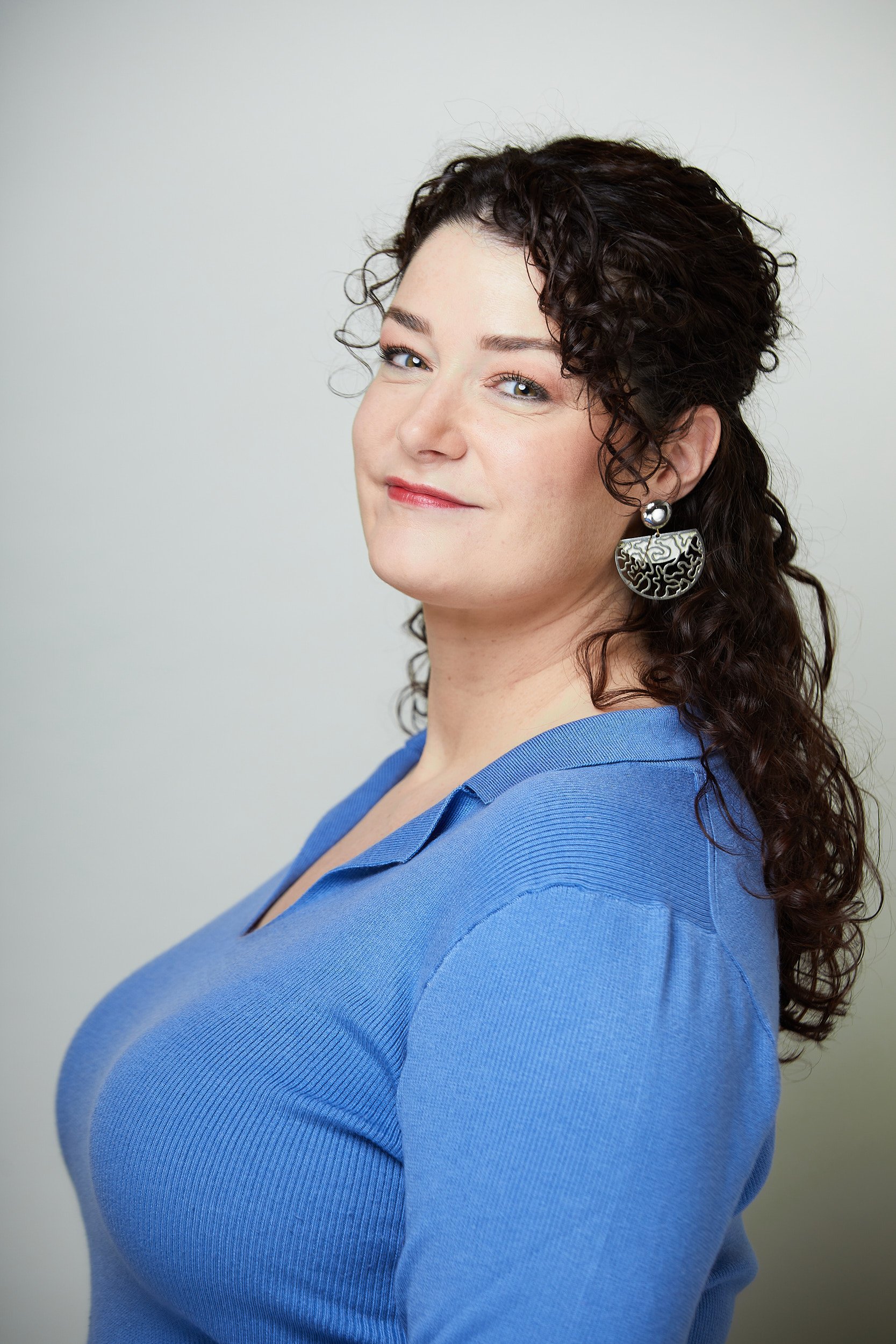 A woman with curly dark hair, wearing a blue top and large earrings, smiling at the camera against a plain background.
