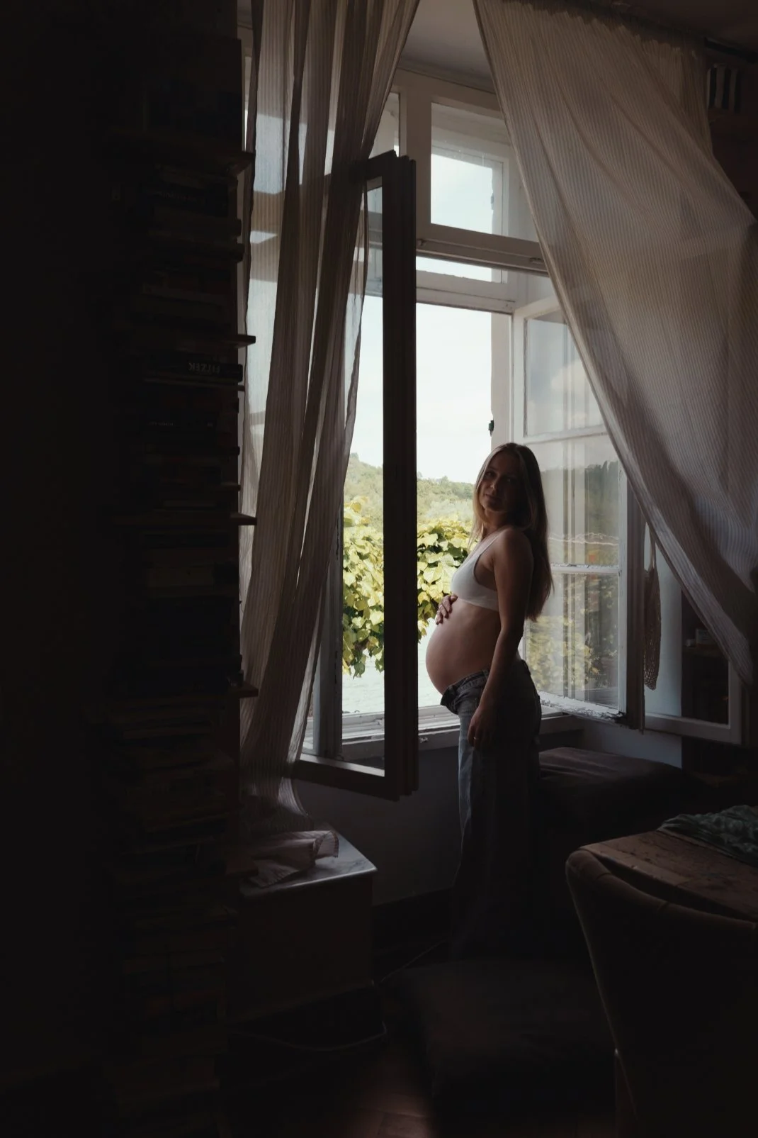 Eine schwangere Frau steht vor einem offenen Fenster, das den Blick auf Bäume und Himmel freigibt. Sie trägt eine weiße Sport-BH und Jeans, und ist im Schatten mit Sonnenlicht im Hintergrund. Das Zimmer hat einen Bücherstapel an der Seite und ein Sof