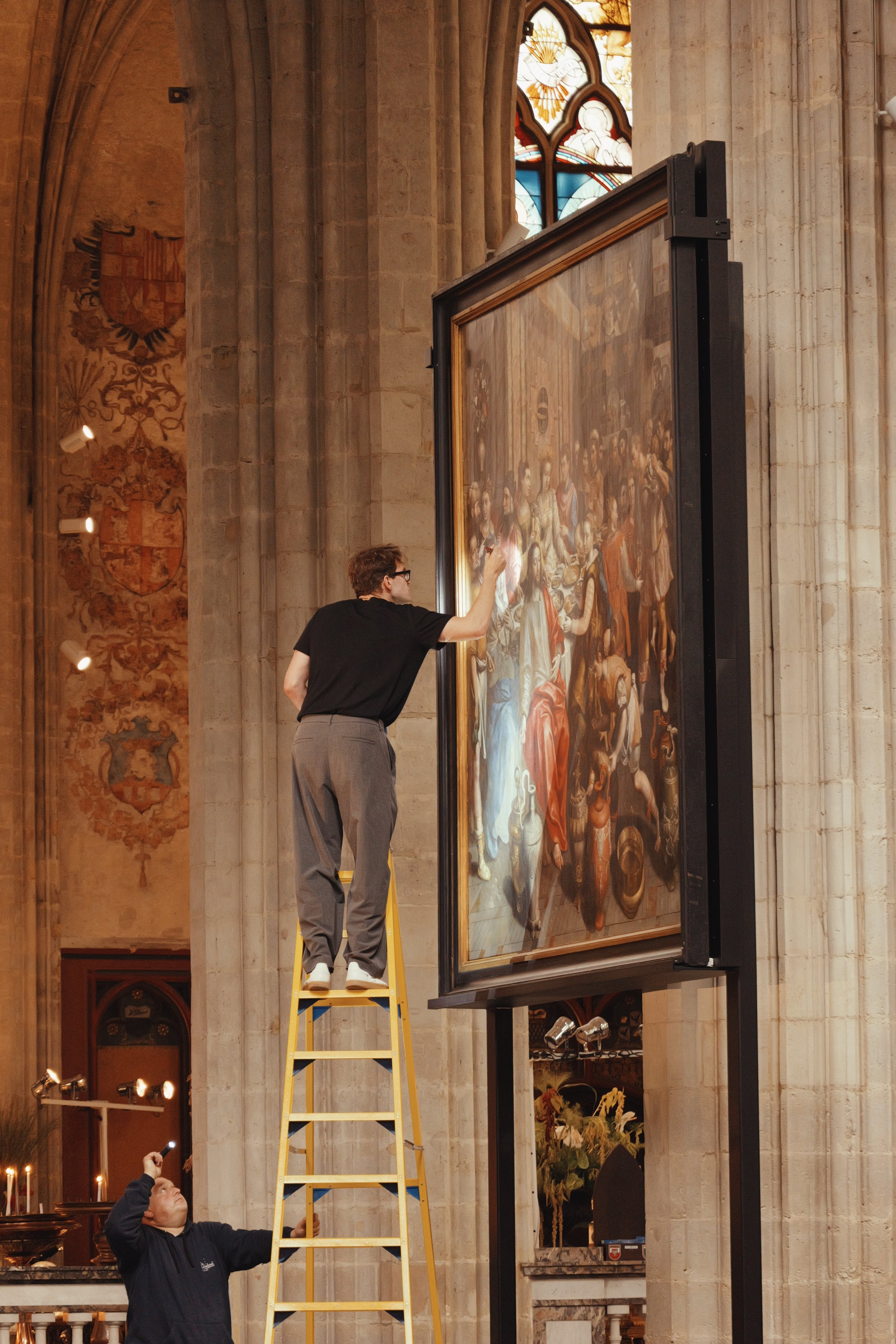 Zwei Männer hängen ein großes Gemälde in einer Kirche auf. Einer steht auf einer Leiter, während der andere unten steht.