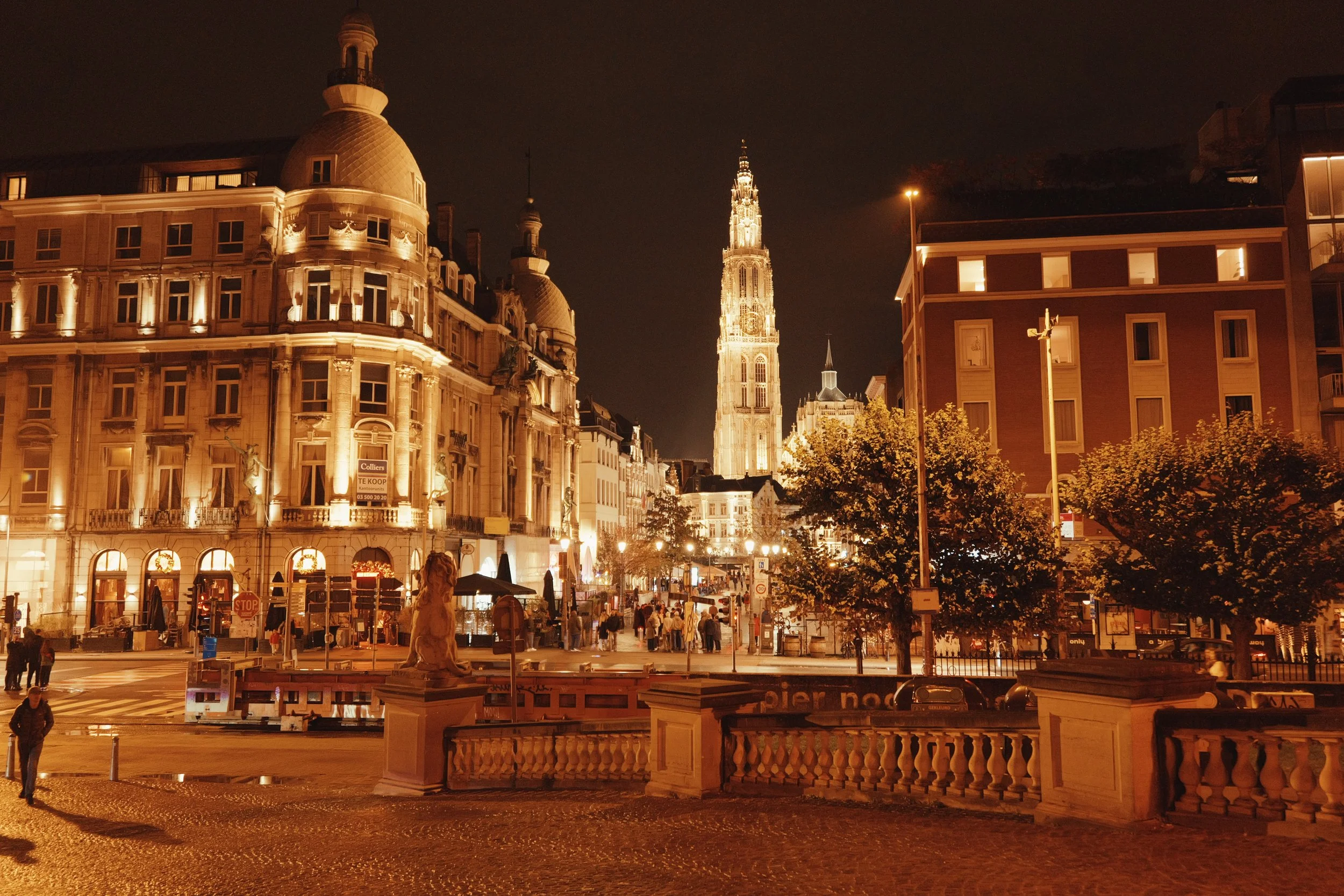 Ein abendliches Stadtbild mit beleuchteten historischen Gebäuden, einer Kirchturm im Hintergrund, Menschen auf belebter Straße und einer Statue im Vordergrund.