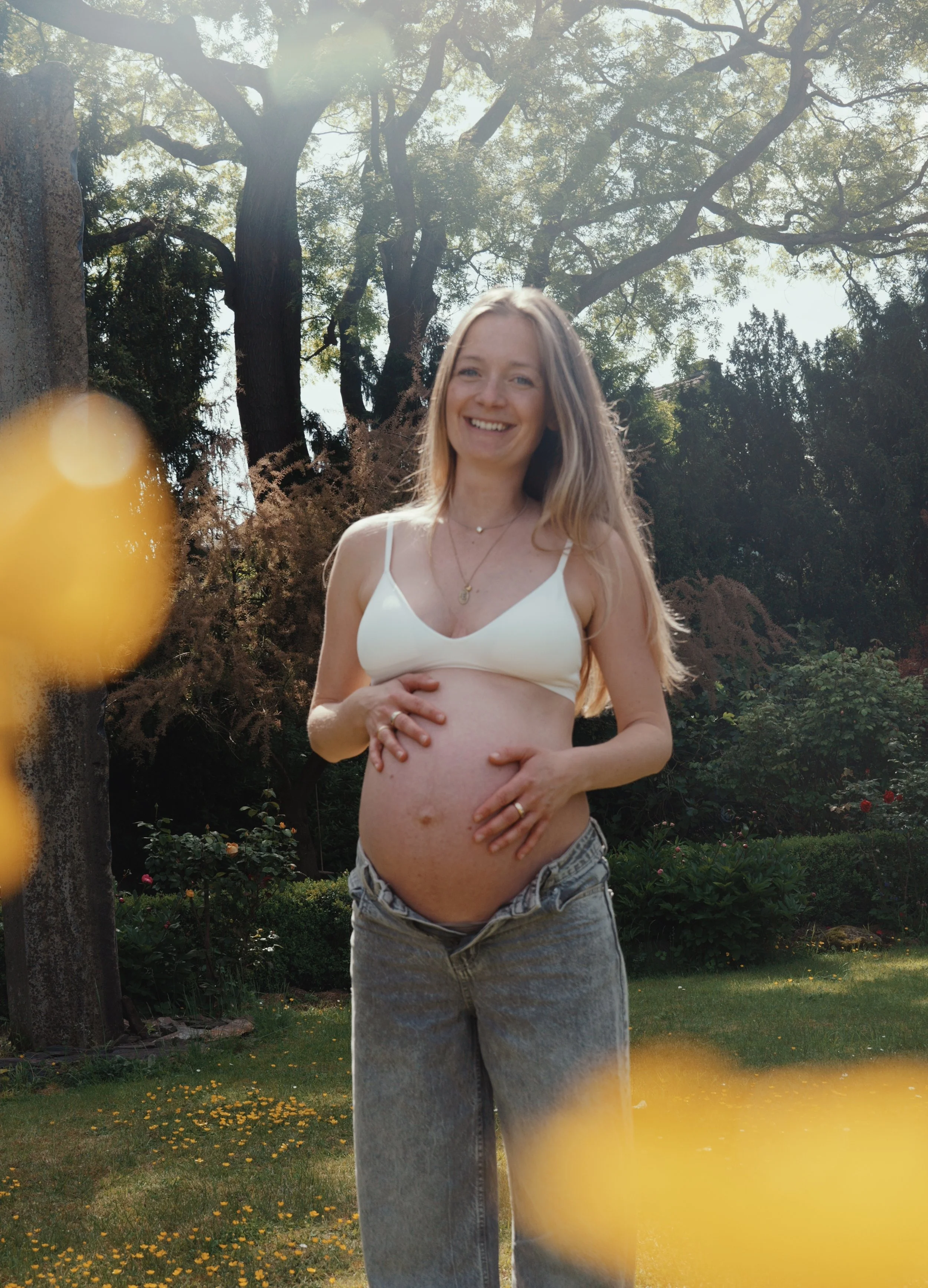 Junge schwangere Frau mit langen Haaren in einem Garten, die ihr Baby hält und lächelnd in die Kamera schaut, umgeben von Bäumen und Blumen, sonniger Tag.