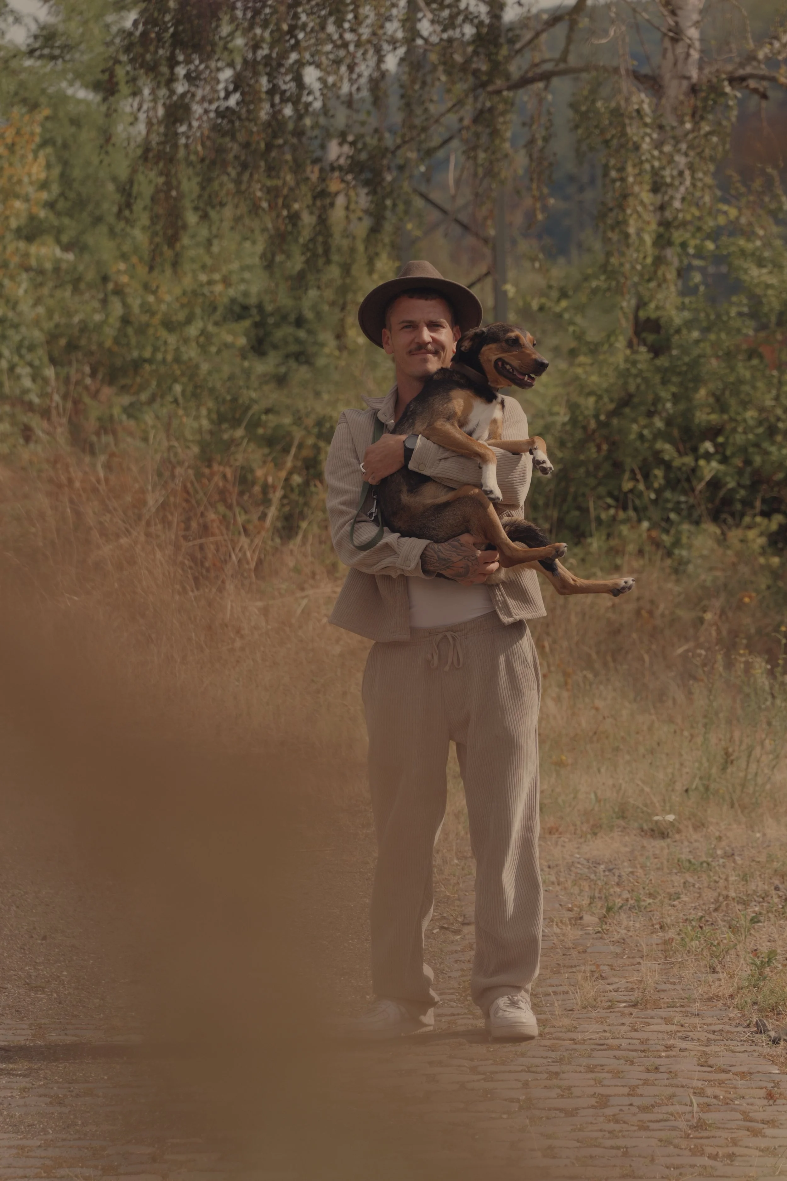 Junger Mann im lässigen Outfit hält lachenden Hund in den Armen beim Spaziergang in einem Park mit Bäumen im Hintergrund.
