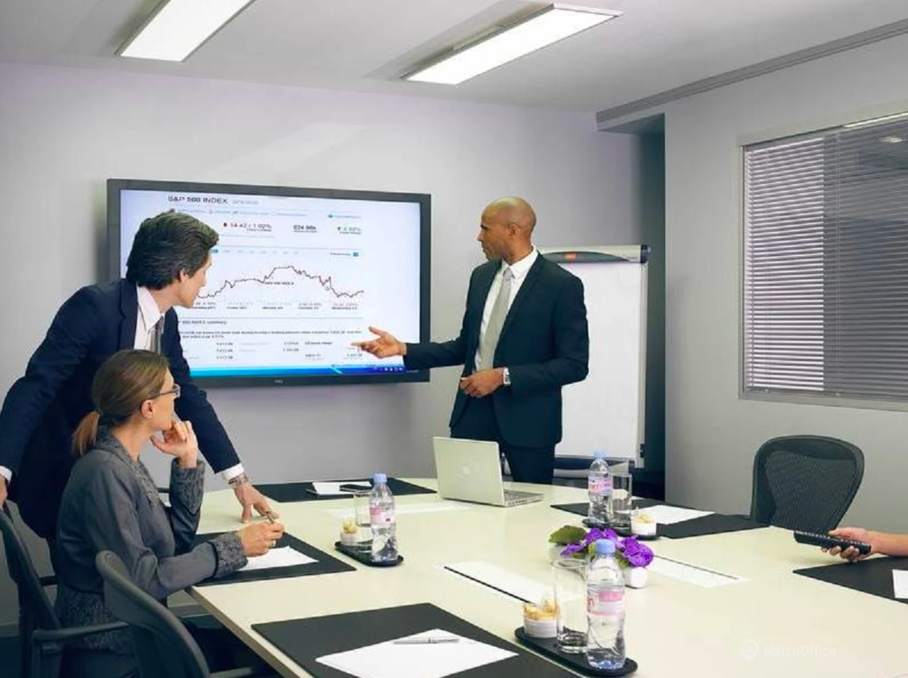 Une réunion d'affaires dans une salle de conférence avec quatre personnes, un homme en costume présente un graphique sur un écran numérique.