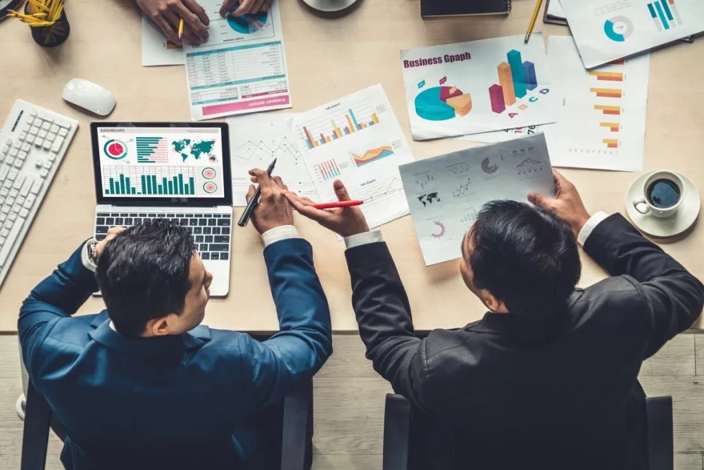 Deux hommes d'affaires debout autour d'une table de réunion examinant des graphiques et des données numériques sur un ordinateur portable et des documents, avec un café sur la table.