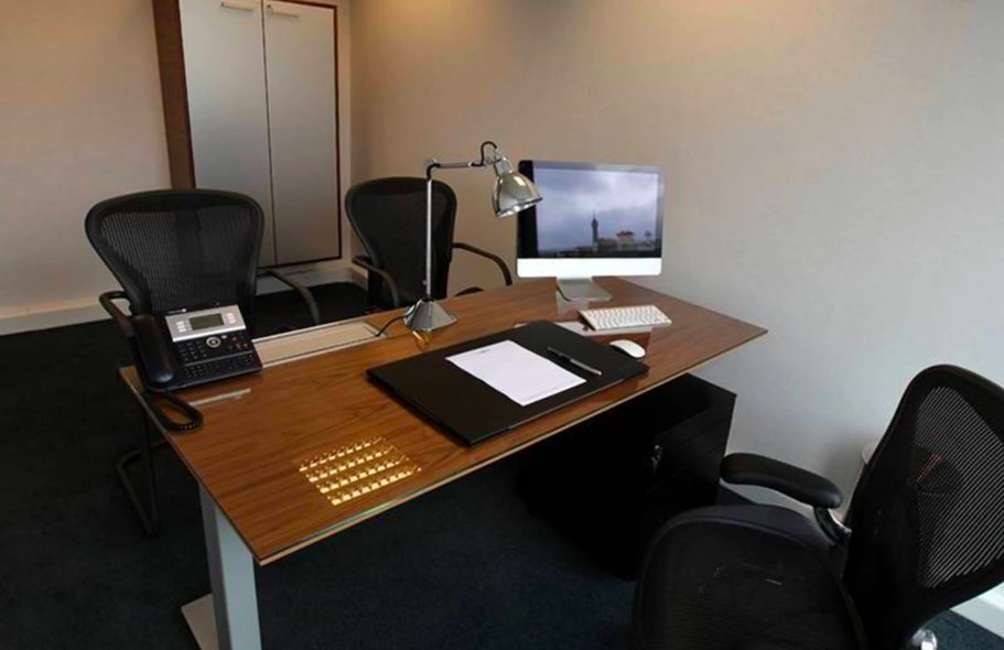 Un bureau avec une grande table en bois, un ordinateur, deux chaises, un téléphone fixe, une lampe de bureau, un clavier, une souris, des papiers, un classeur, un fichier noir, et un mur blanc