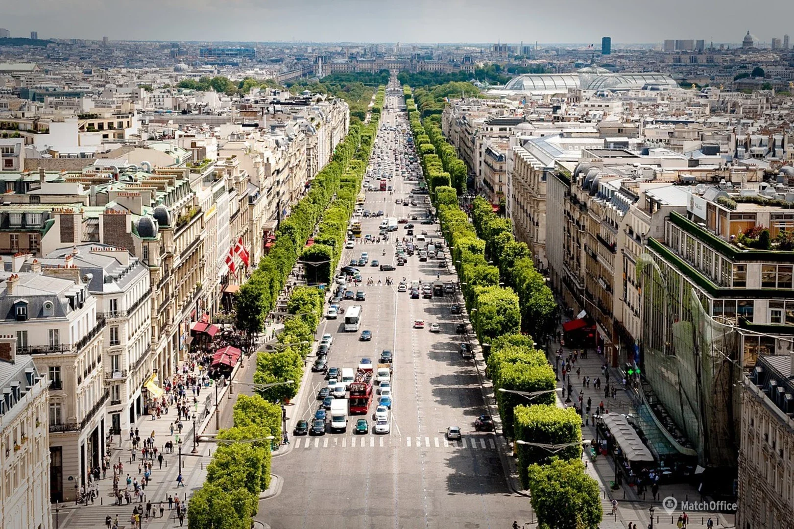 Vue aérienne d'une avenue animée bordée d'arbres verts, avec des bâtiments historiques et modernes, des véhicules et beaucoup de piétons, sous un ciel partiellement nuageux à Paris.