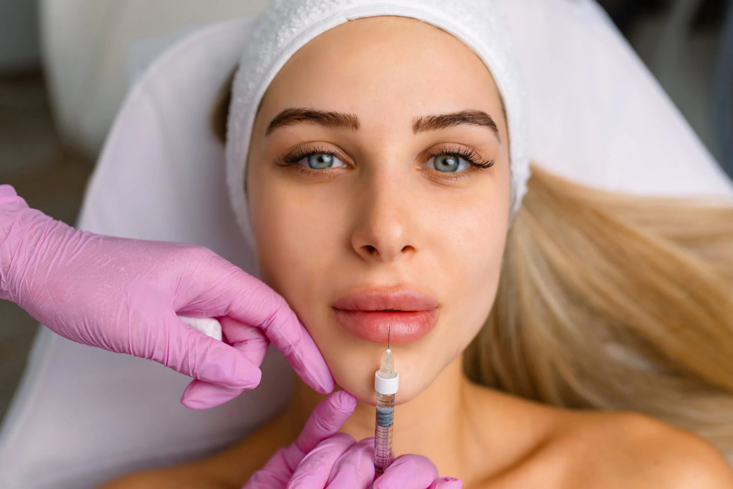 A woman with blue eyes and light skin receives an injecton in her chin area from a medical professional wearing pink gloves, while lying on a treatment bed.