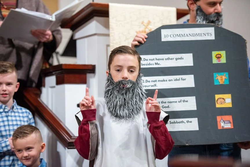 Boy dressed as Moses with a gray beard, pointing upward, in front of a blackboard displaying the 10 Commandments with small colorful icons.