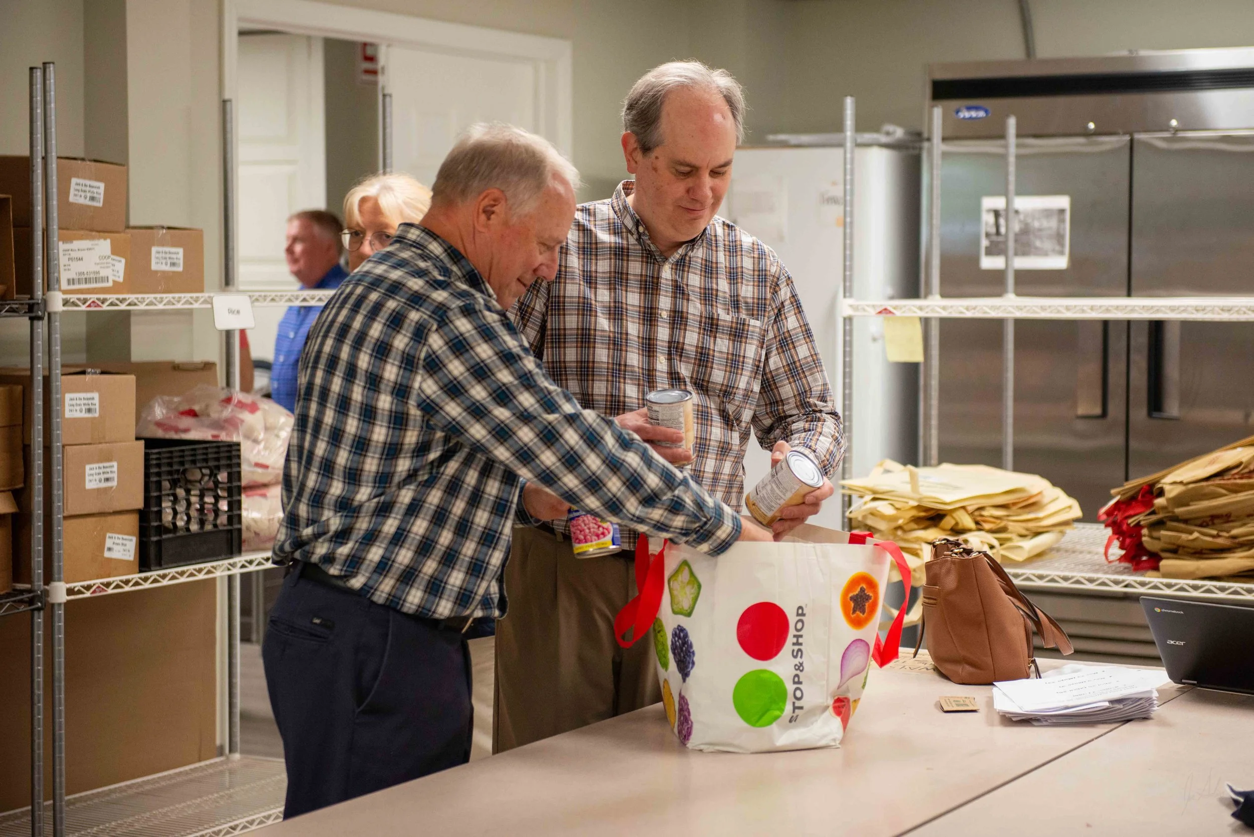 Two men in checkered shirts packing food items at a table in a storage or distribution room with metal shelves filled with boxes and packages.