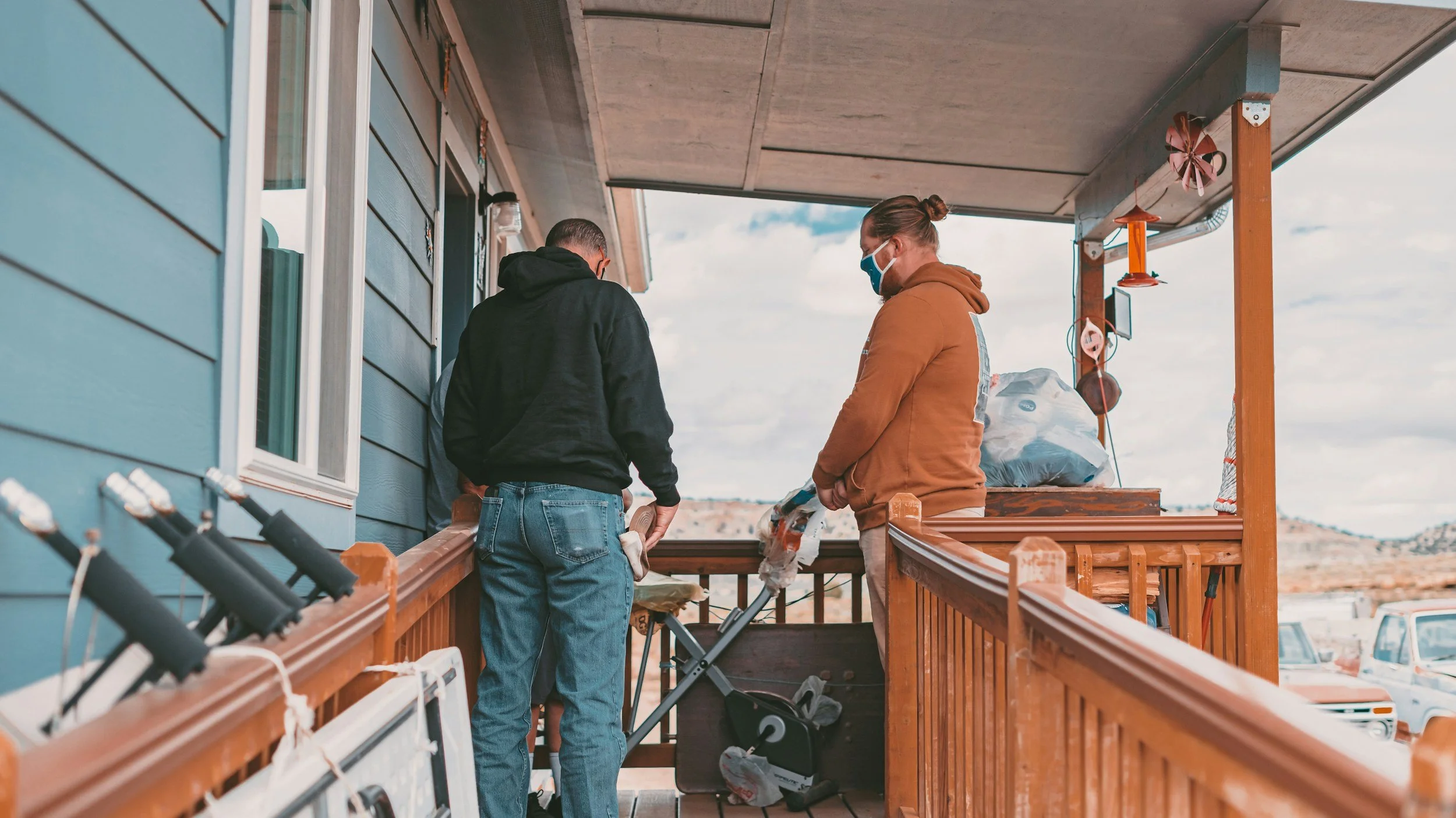 Two people wearing masks stand on a wooden porch, working on a project with tools and materials nearby. The house has blue siding, and the sky is partly cloudy.
