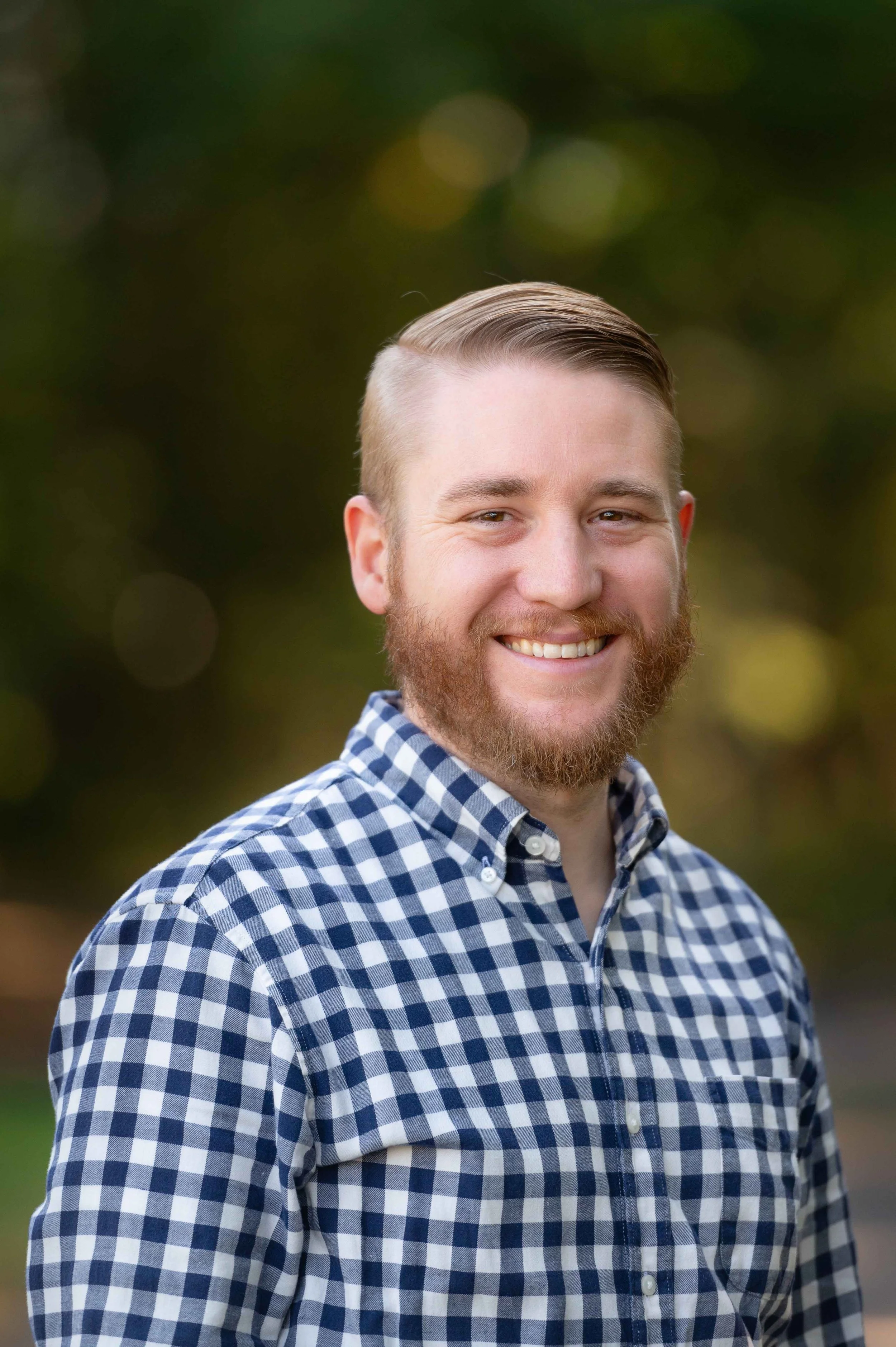 A smiling man with a beard and slicked-back hair wearing a blue and white checkered shirt outdoors with a blurred green background.