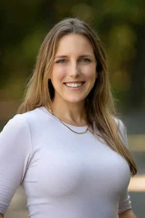 A woman with long light brown hair smiling outdoors with trees in the background, wearing a white top and a gold necklace.